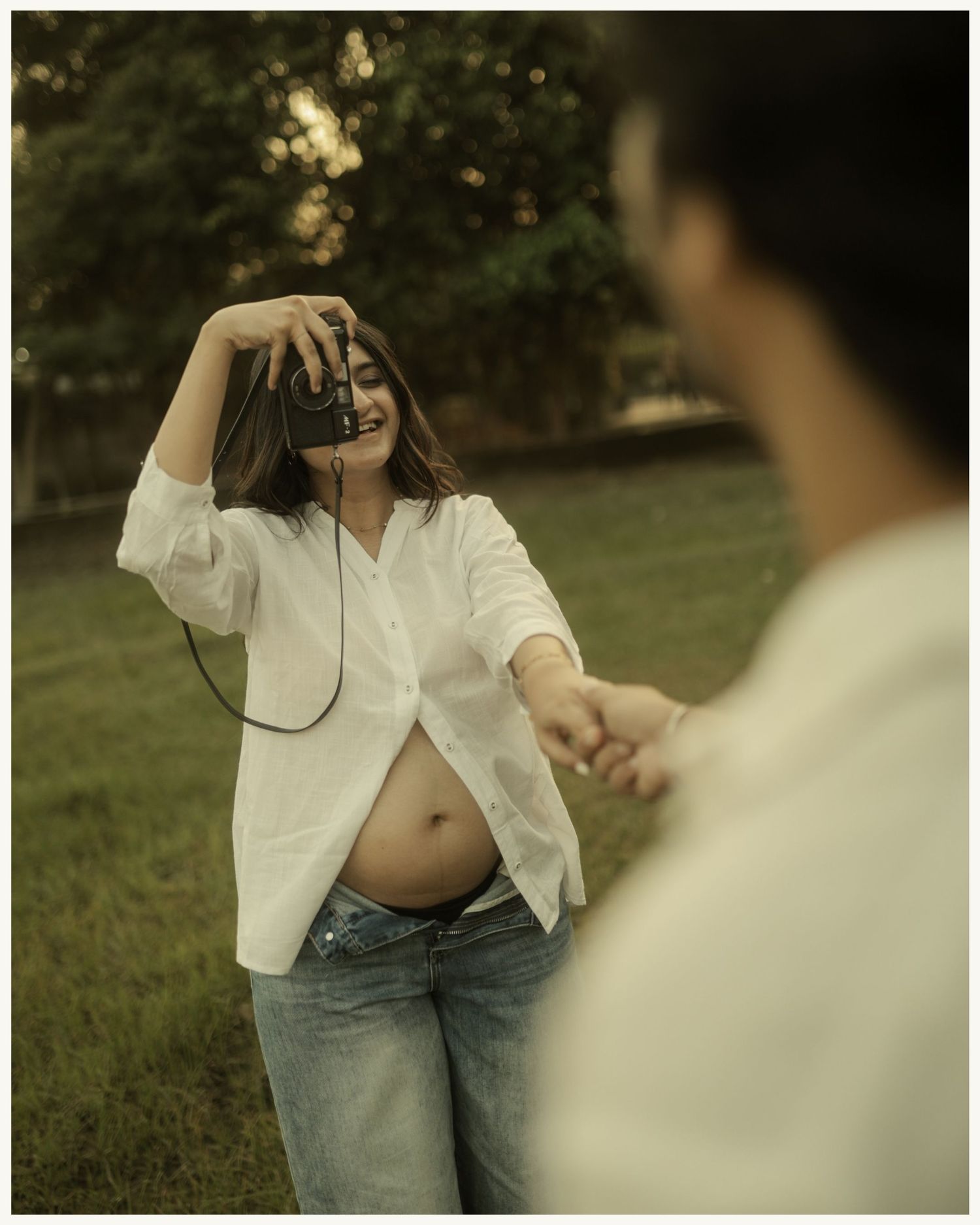 Smiling pregnant woman in white shirt and jeans holds vintage camera outdoors while partner holds her hand.
