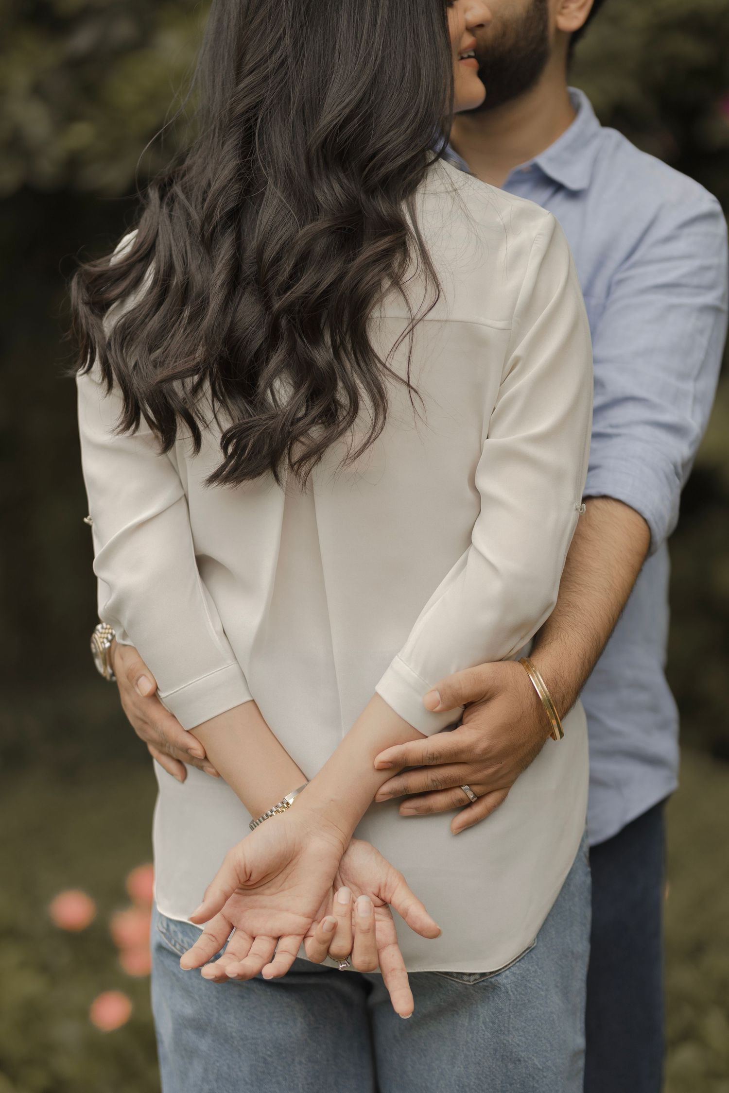 Romantic couple embracing from behind, holding hands, woman in white blouse with wavy hair, man in blue shirt outdoors.