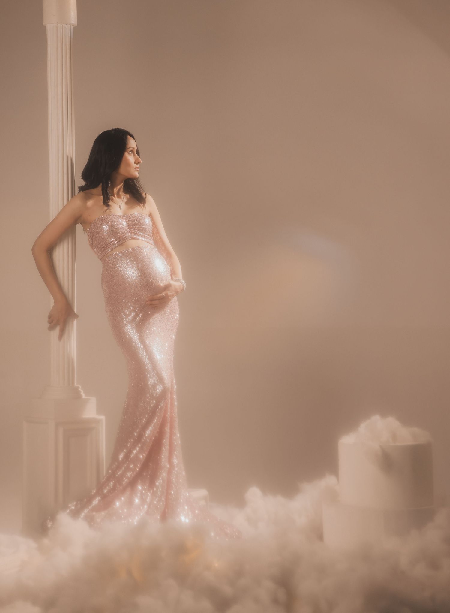 Pregnant woman in a pink sequin gown poses elegantly by a white column, surrounded by soft clouds in a dreamy setting.