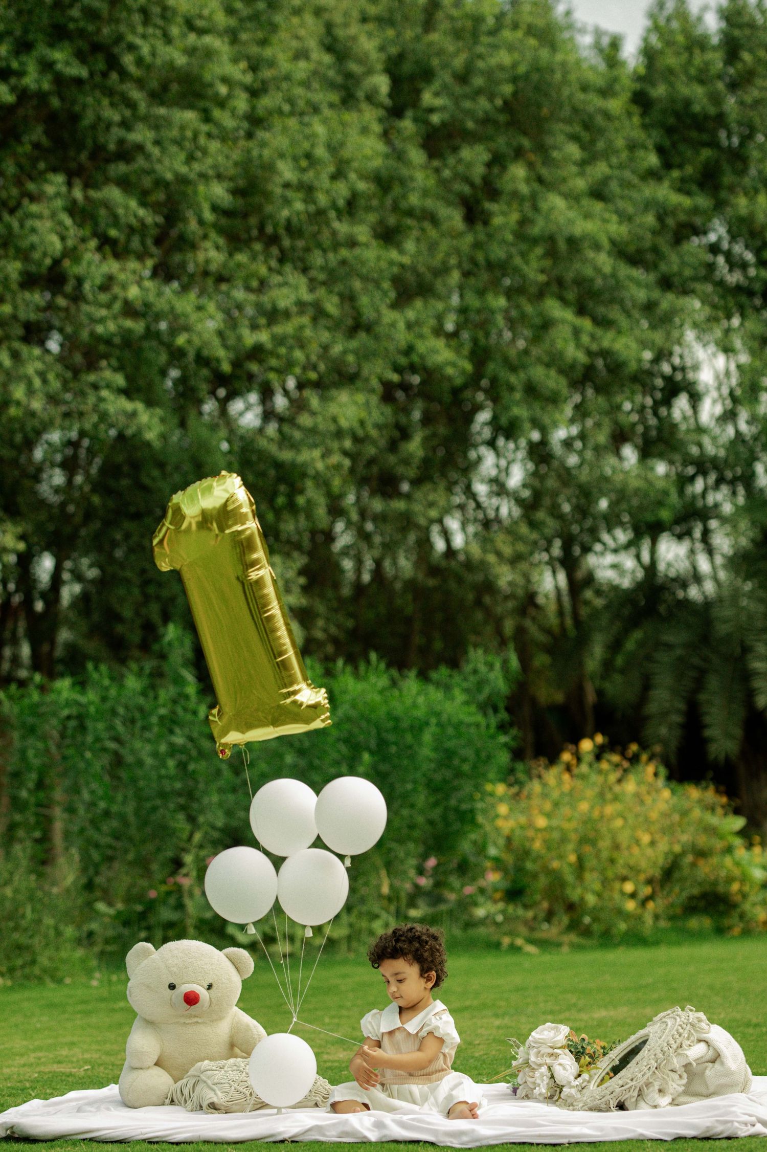 Baby's first birthday outdoor celebration with gold number one balloon, white teddy bear, and balloons on a green lawn.