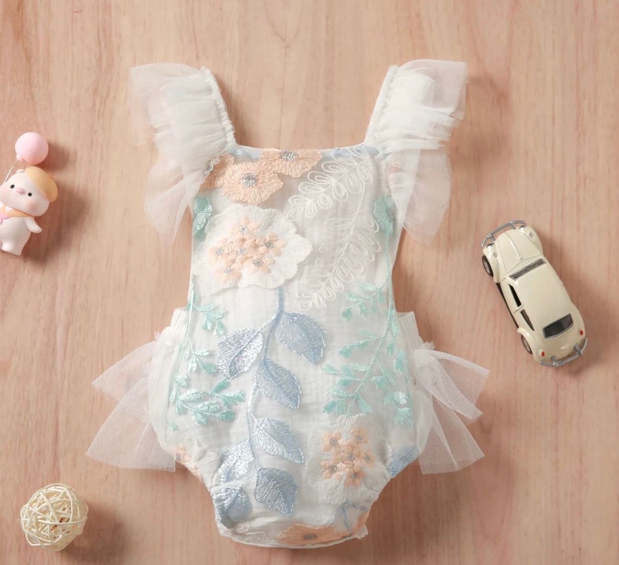 A delicate floral print baby romper with tulle accents displayed on wooden surface next to small toy and miniature car.
