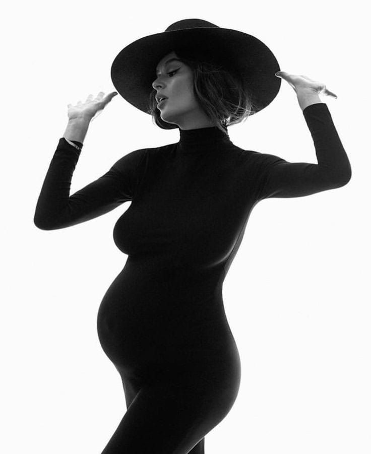 Striking silhouette of pregnant figure in black turtleneck and wide-brimmed hat.