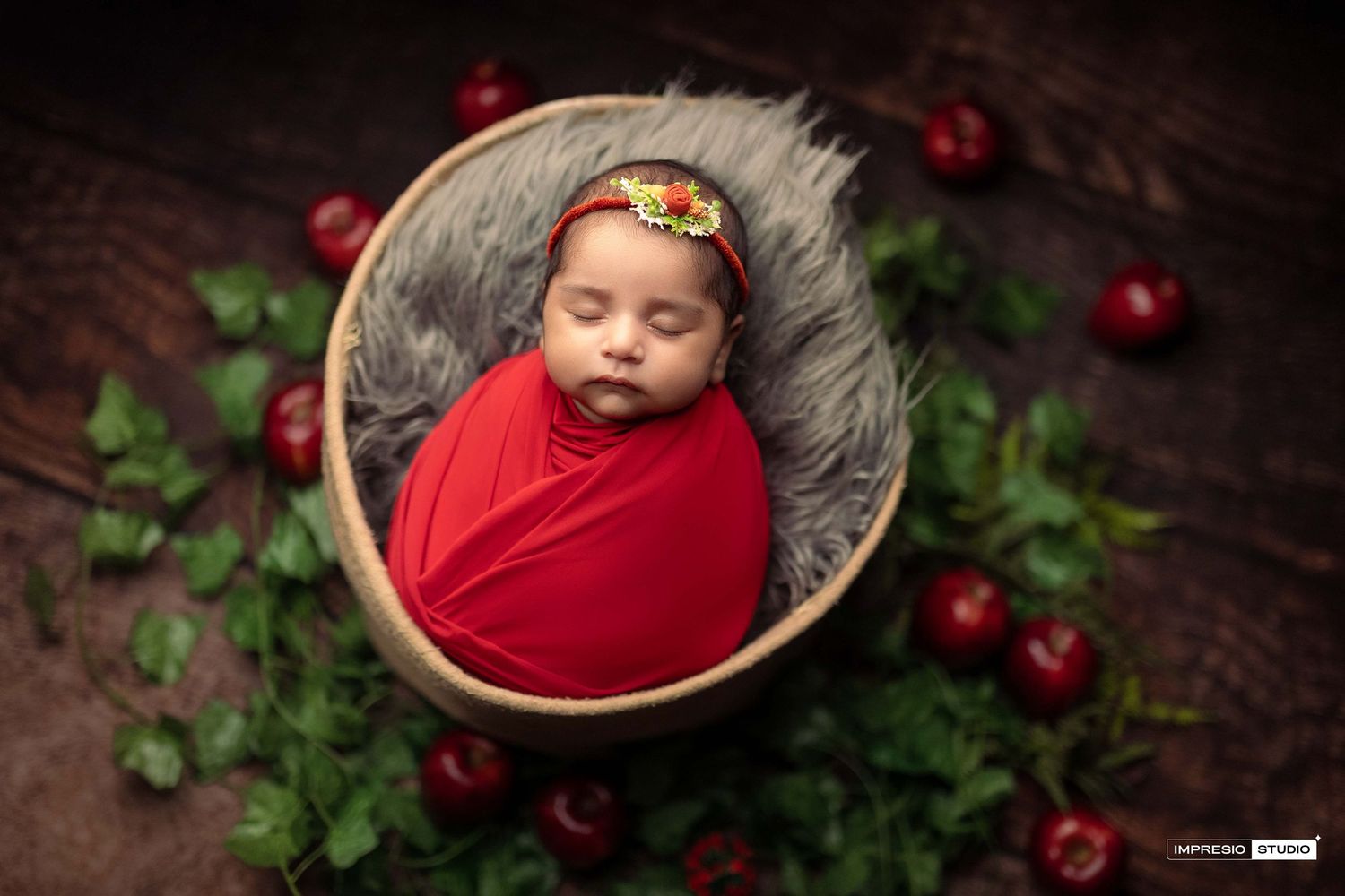 Newborn baby wrapped in red swaddle sleeping in wooden bowl with gray fur, flower headband, and red apples around it.