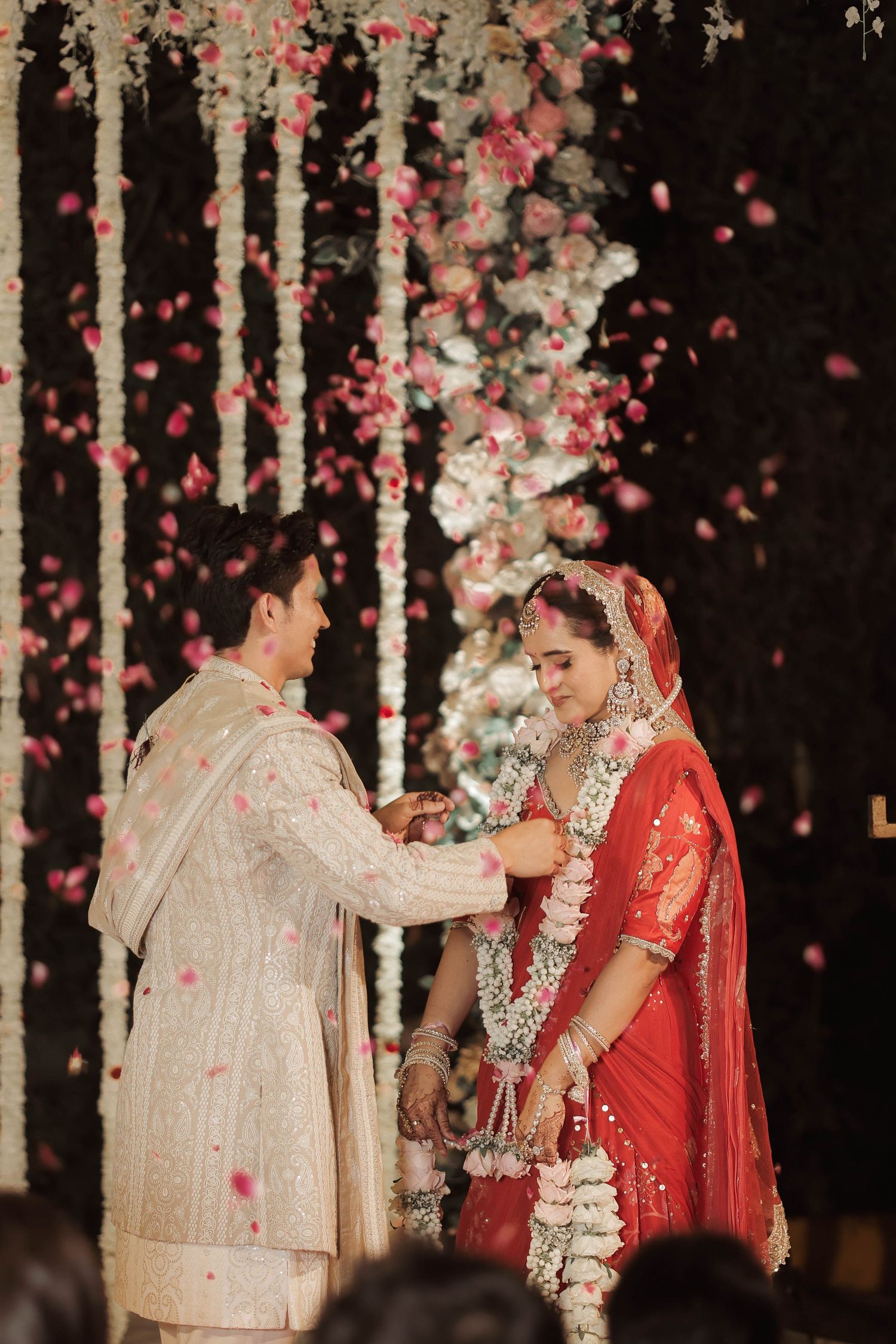 Indian bride in red saree and groom in white sherwani exchange garlands at wedding ceremony with flower petals falling.