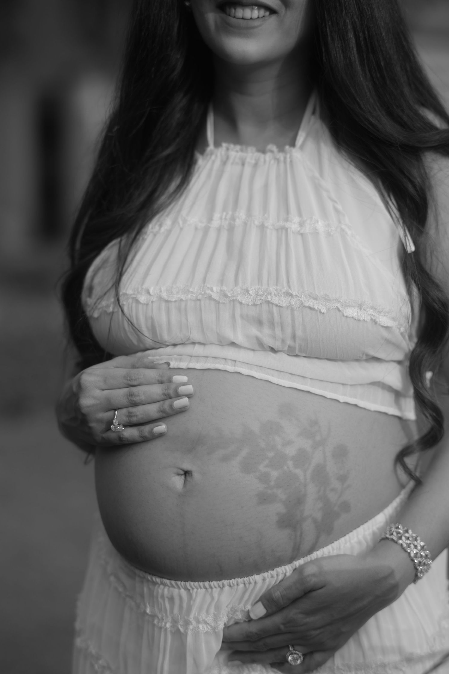 A black and white maternity photo series showing a pregnant belly in a white ruffled top with hands gently cradling it.