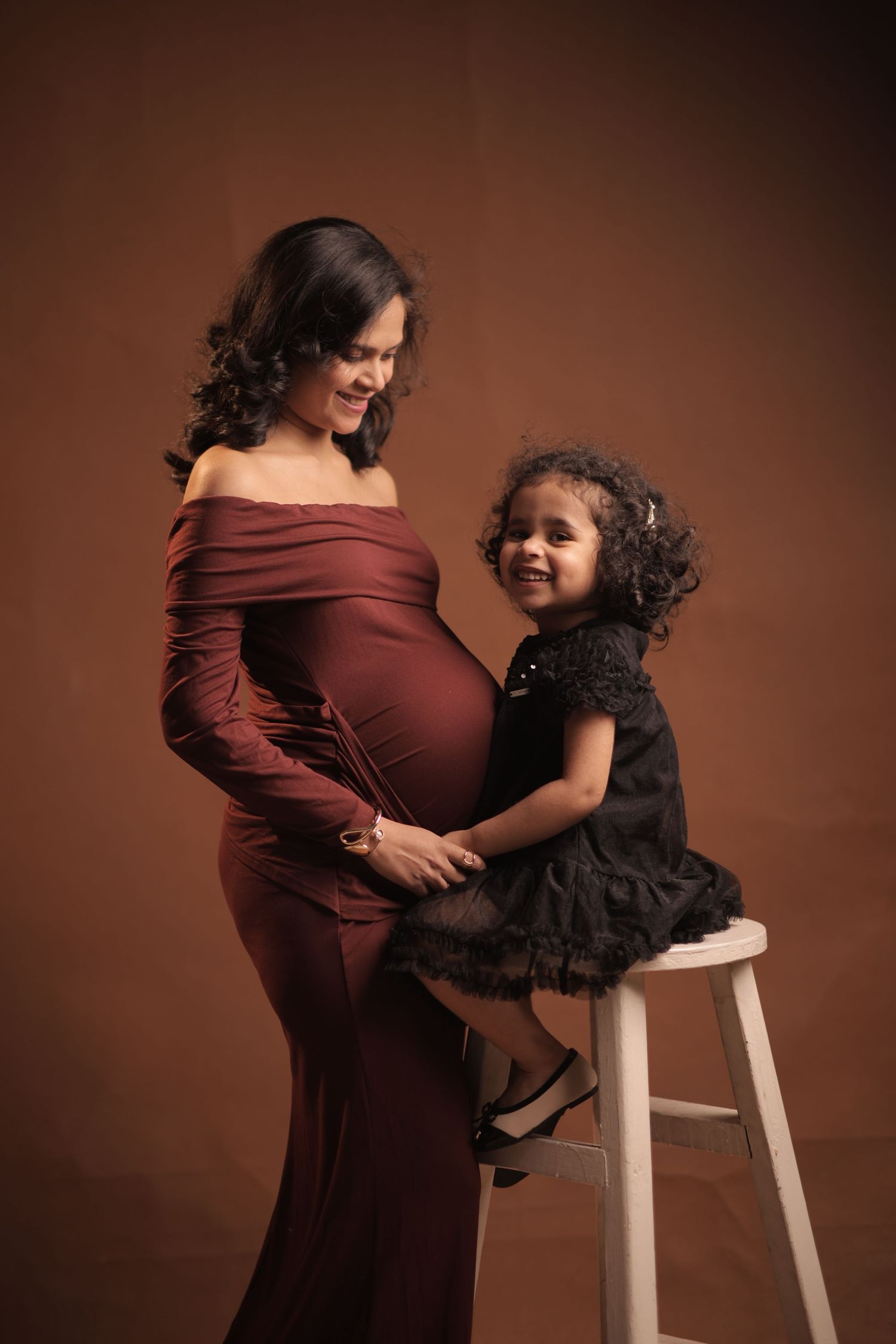 A maternity photography series shows tender moments between a pregnant woman in burgundy dress and child against brown backdrop.