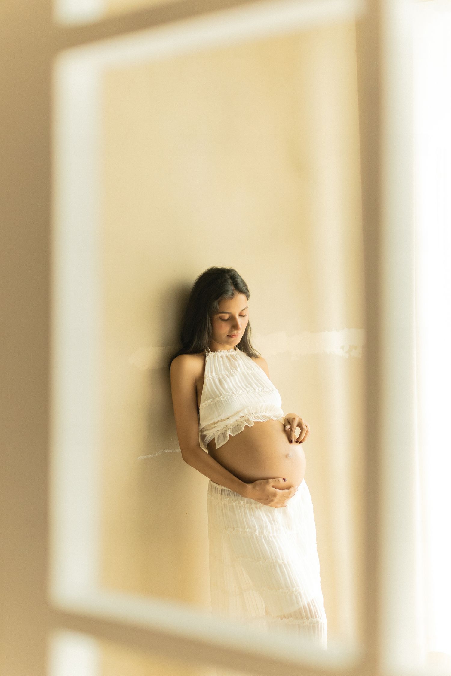 A pregnant woman in a white dress stands by sunlit windows in an ethereal maternity photo sequence.