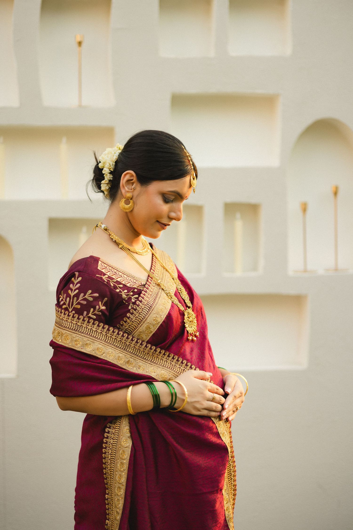 A person wearing a burgundy and gold traditional Indian saree stands against a light-colored wall.