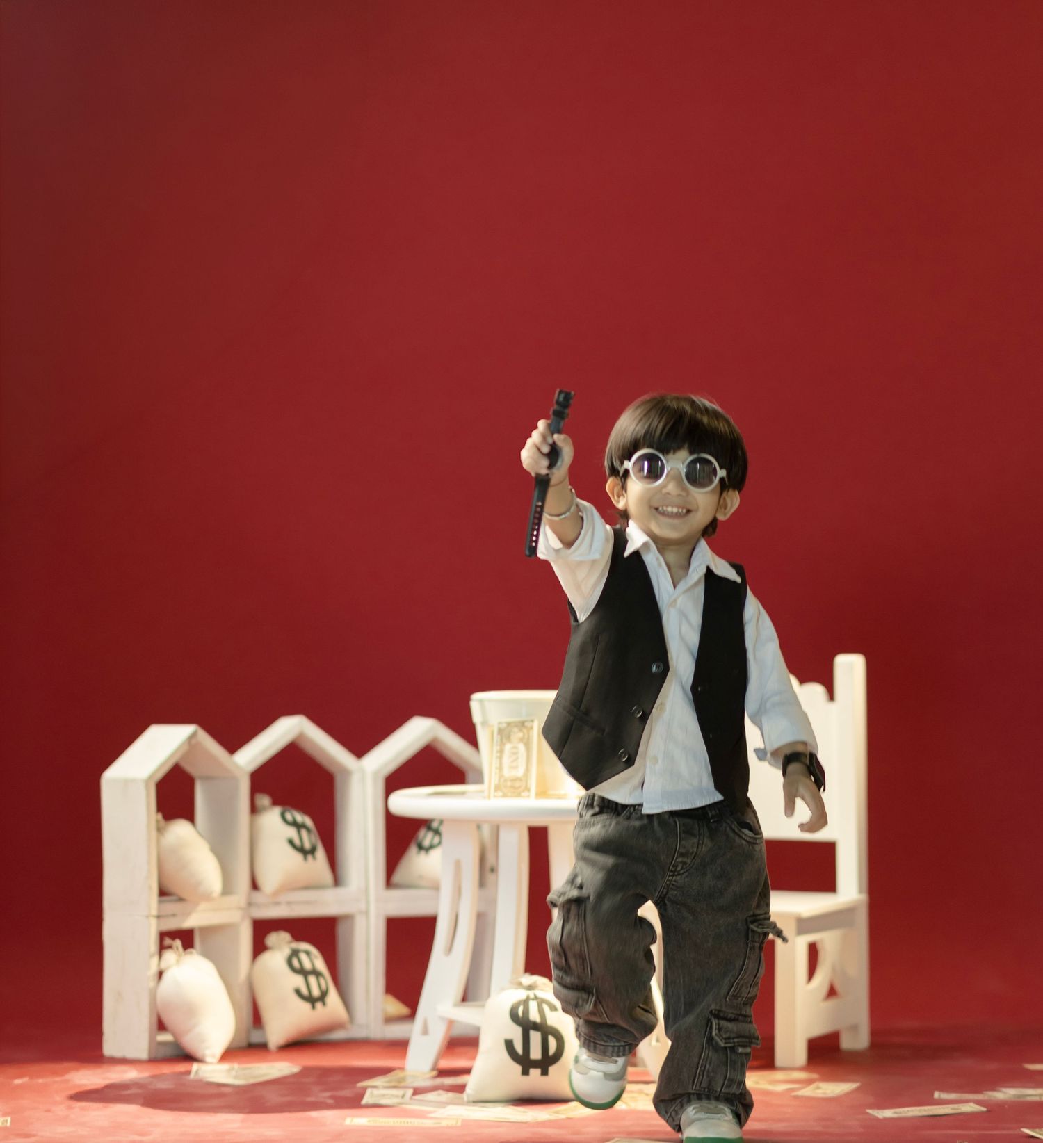 A boy doll wearing sunglasses and a vest poses with money bags and white fence props on a red background.