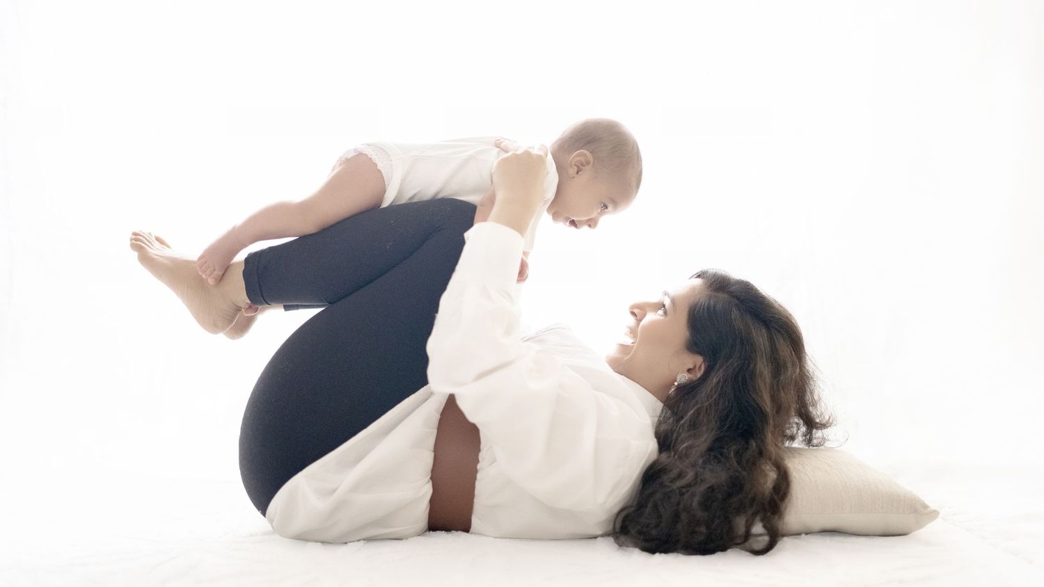 A mother lies on her back lifting her baby in the air during a playful yoga session on a white background.
