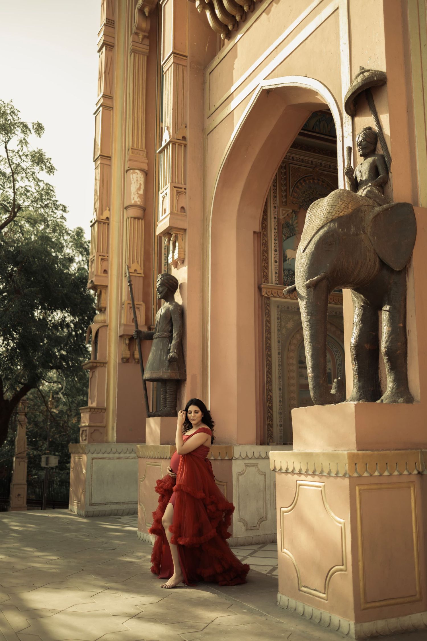 Woman in flowing red dress does an elegant maternity pose next to ornate palace entrance with elephant statues in India.