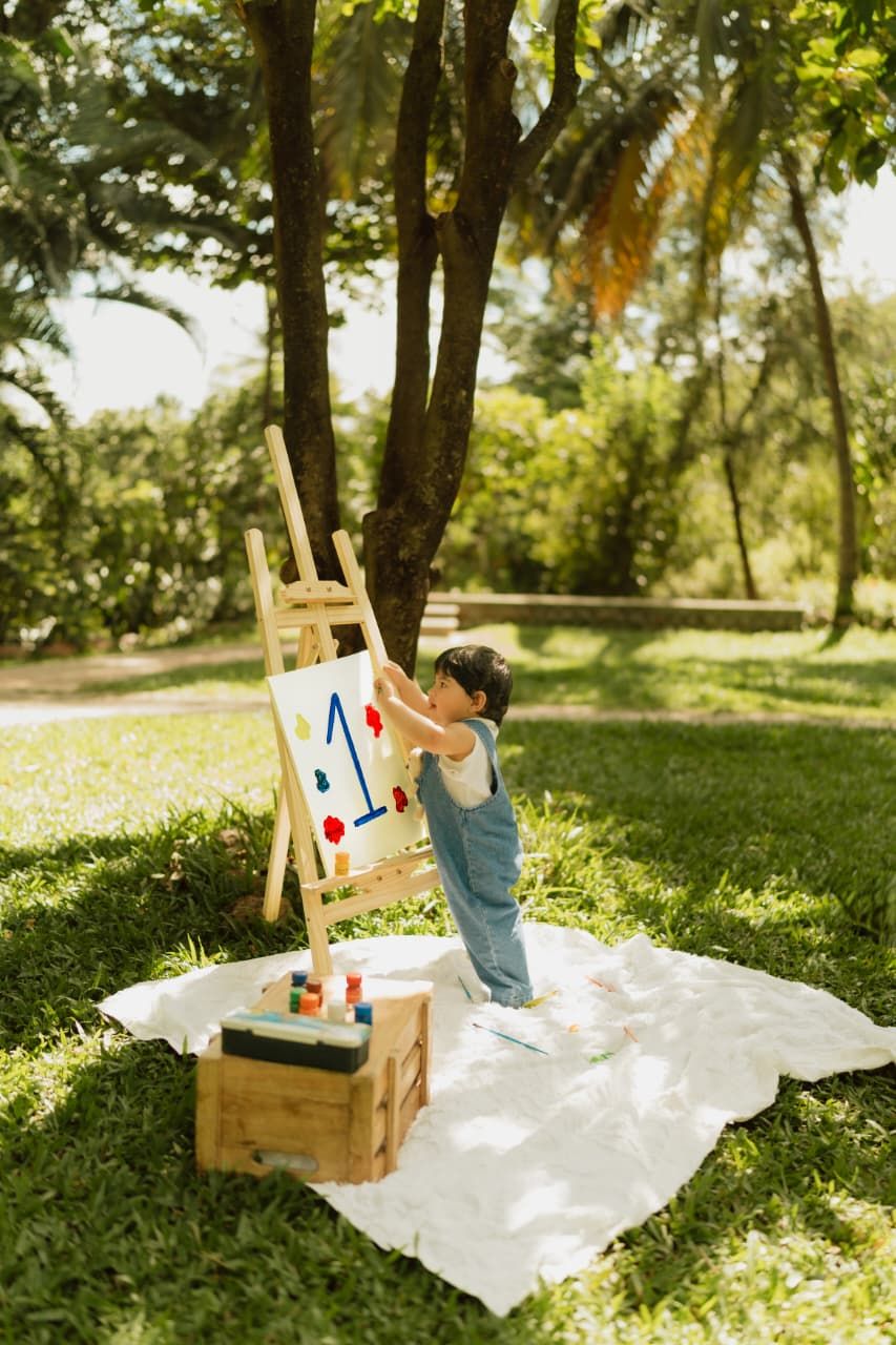 Toddler painting on an easel outdoors in a lush green park, standing on a white blanket with paint supplies nearby.