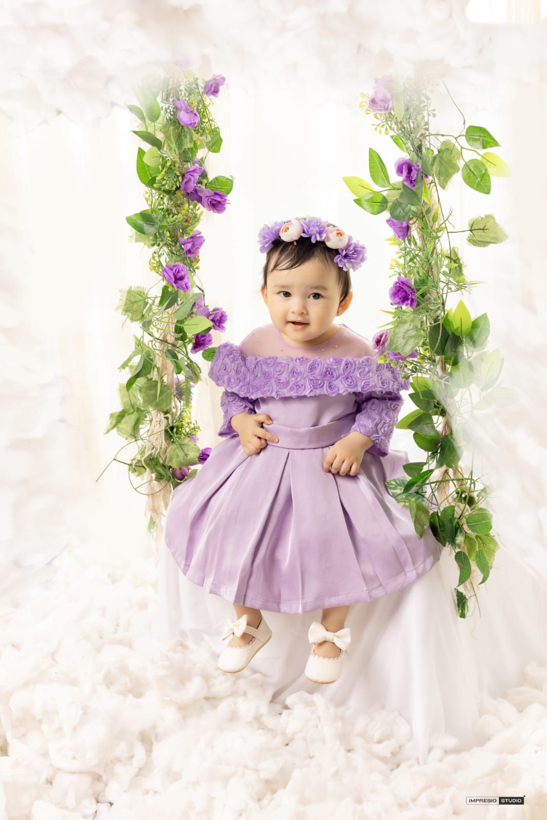 Baby girl in lavender dress and floral crown sits on a flower-adorned swing surrounded by purple roses and green vines.