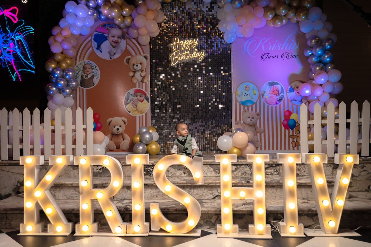 Child sits behind illuminated marquee letters spelling KRISHIV at a colorful birthday party with balloons and teddy bear backdrop.