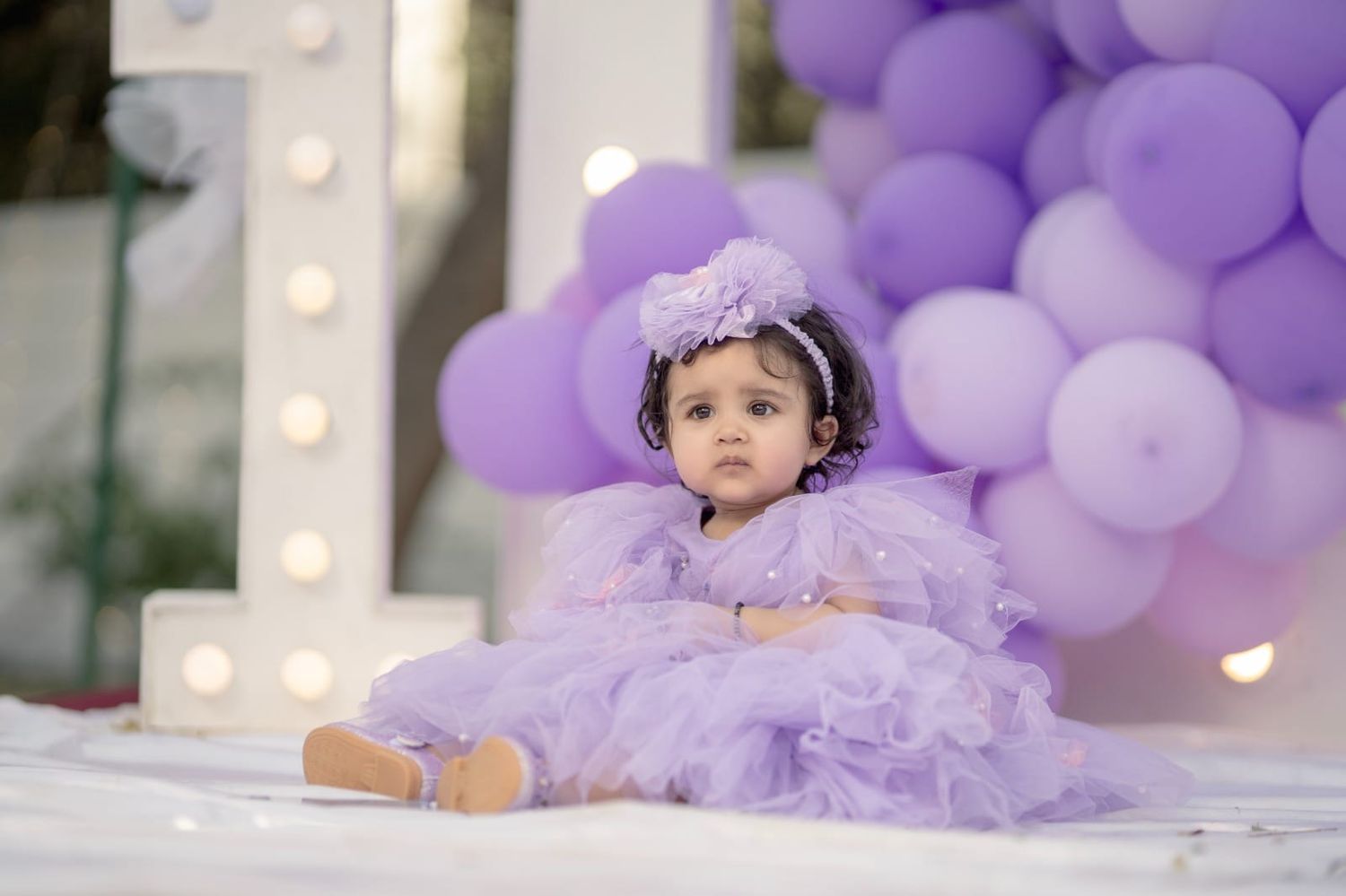 A baby girl in a purple tulle dress sits among lavender balloons at a birthday celebration.