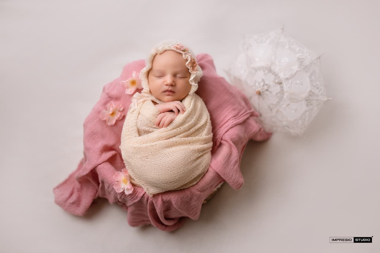 Newborn baby girl swaddled in cream wrap on pink blanket with floral headband and lace decoration.