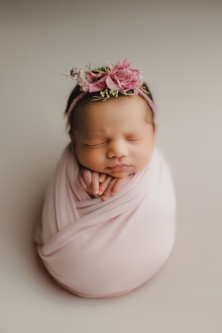 Sleeping newborn baby swaddled in pink wrap with floral headband, peaceful studio portrait photography.