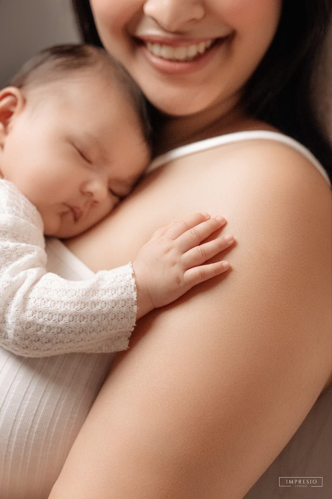 Smiling mother tenderly holds sleeping newborn baby dressed in white against her chest.