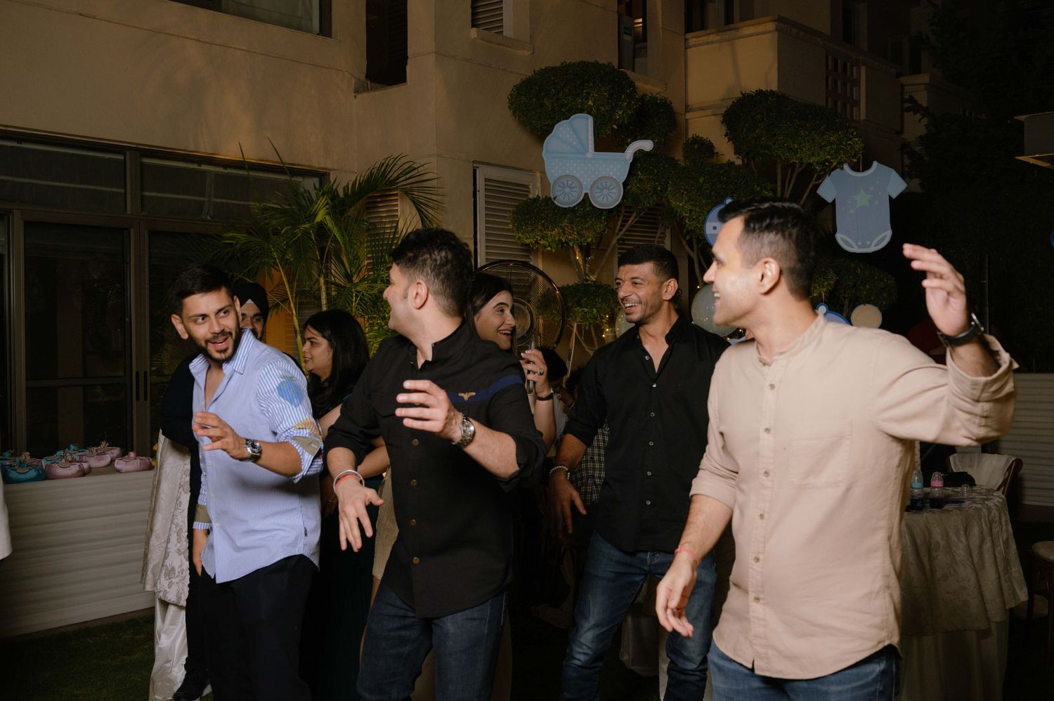 Group of men dancing and celebrating at an outdoor baby shower party at night, holding baby onesie decorations.