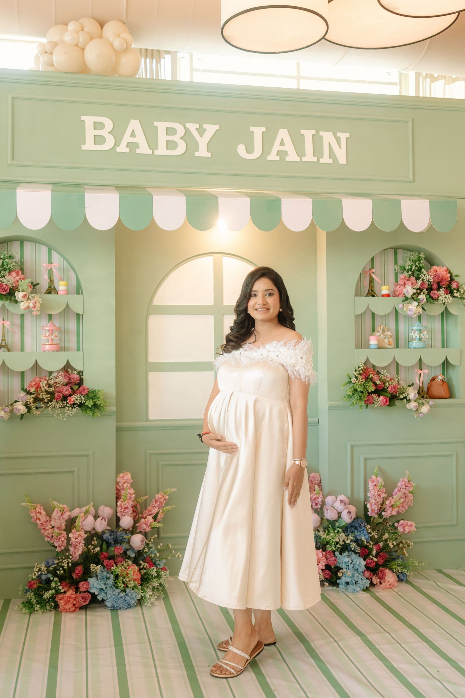Pregnant woman in white off-shoulder dress poses at a mint green Baby Jain baby shower backdrop with floral arrangements.