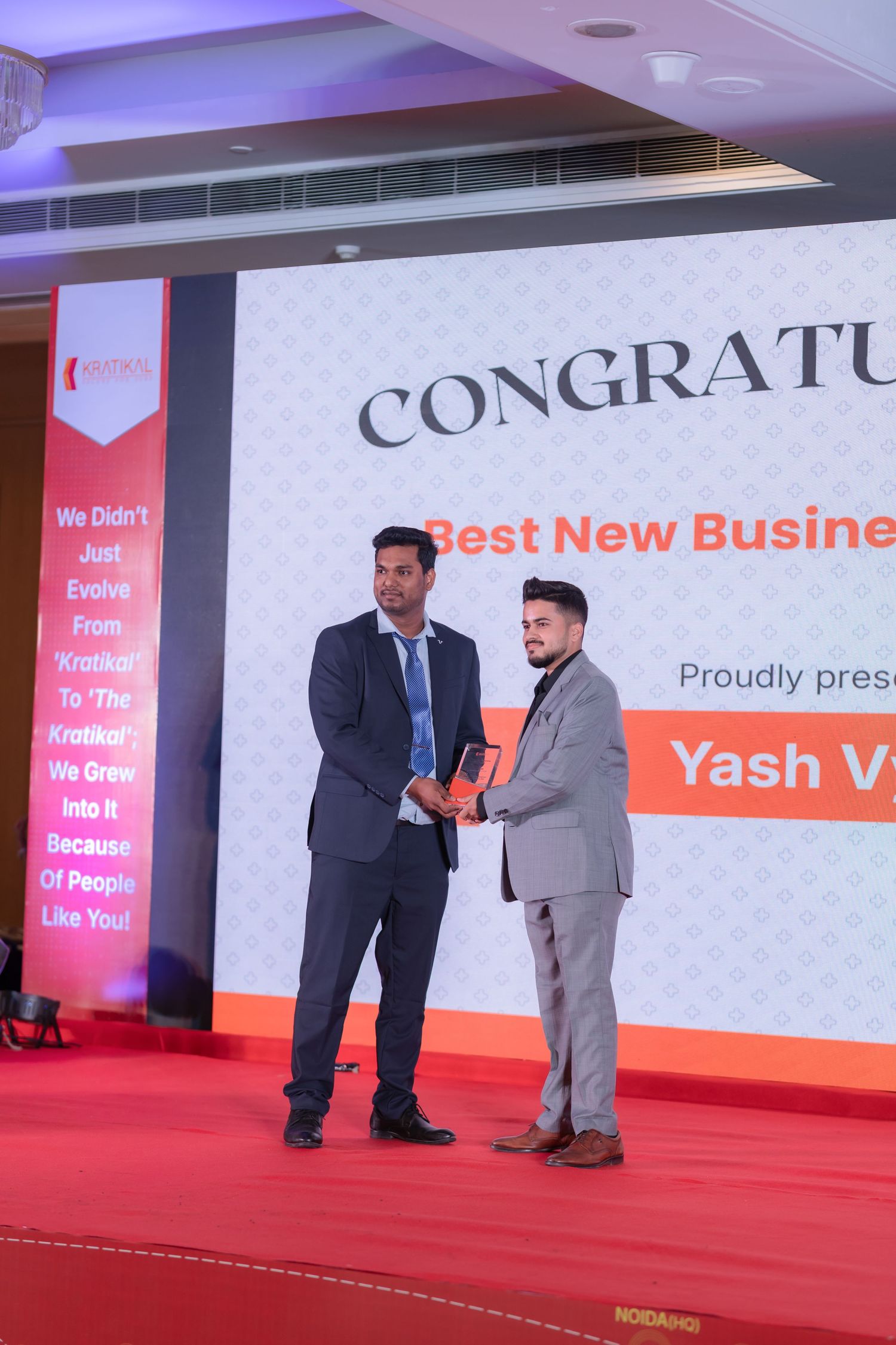 Two men in suits on a red stage exchange a trophy at a Kratikal awards ceremony celebrating the Best New Business award.