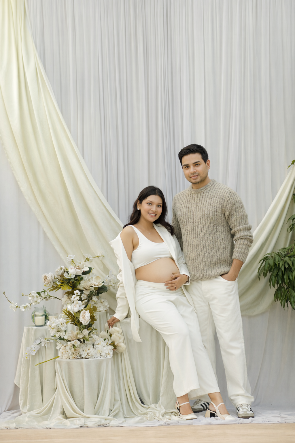 A couple's maternity photoshoot with white drapes, floral decor, and coordinated neutral outfits.