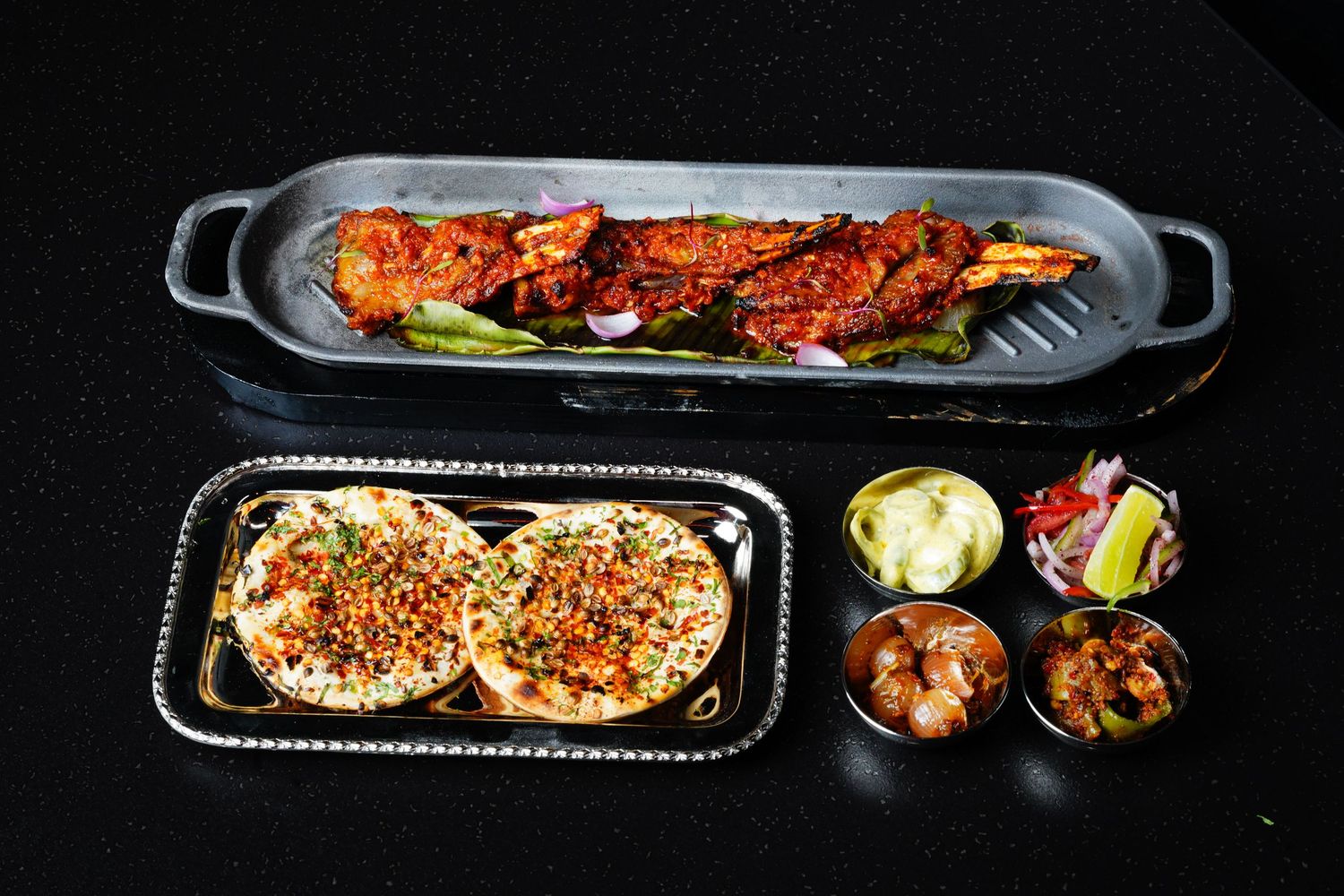 Various grilled and seasoned dishes displayed in black serving trays against a dark background.