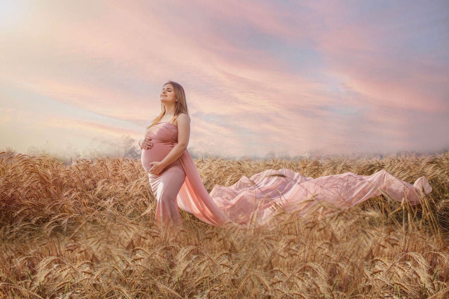 Serene maternity session in a golden wheat field during sunset with flowing pink dress.