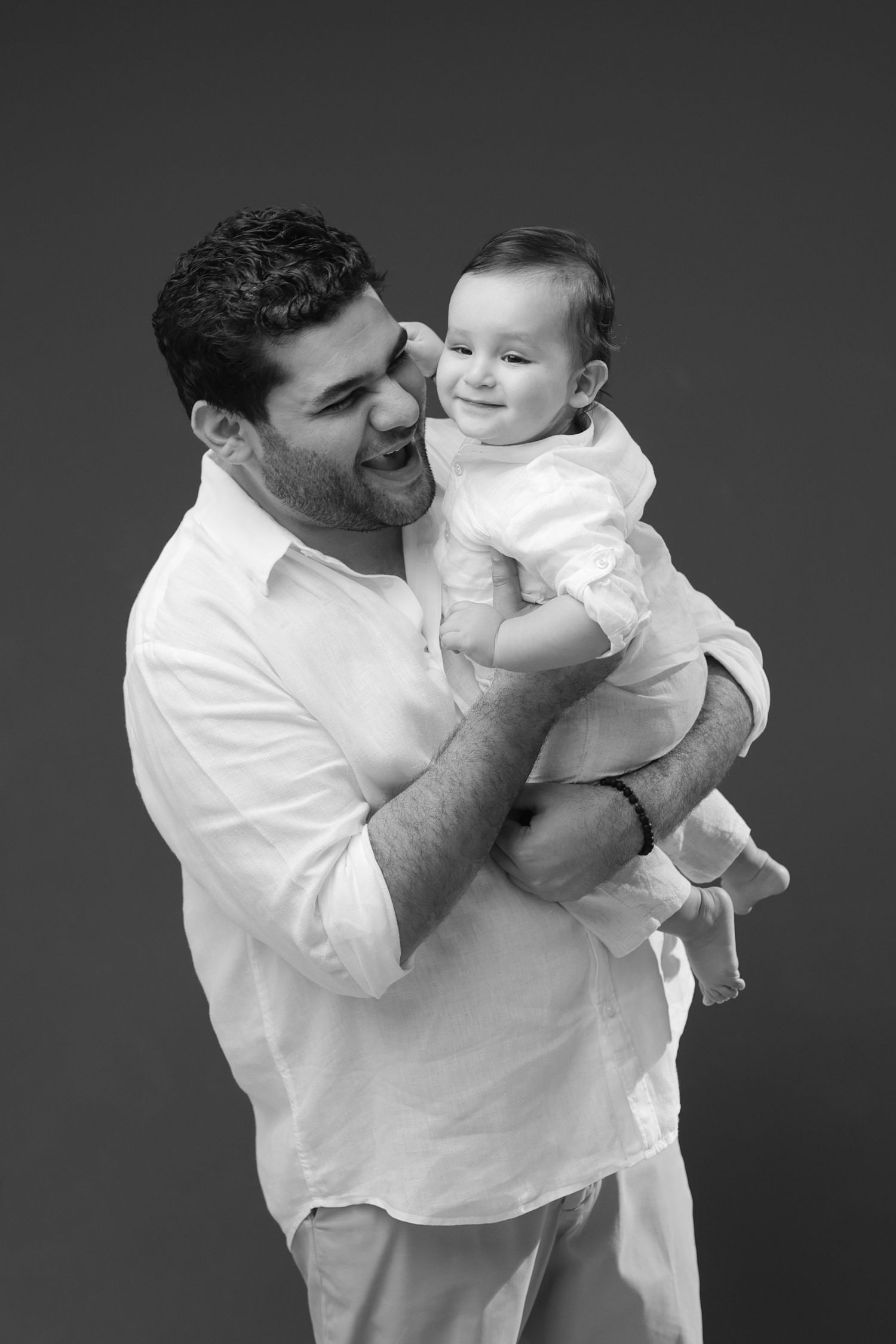 Black and white studio photo of a smiling man in white shirt holding a happy baby dressed in white.