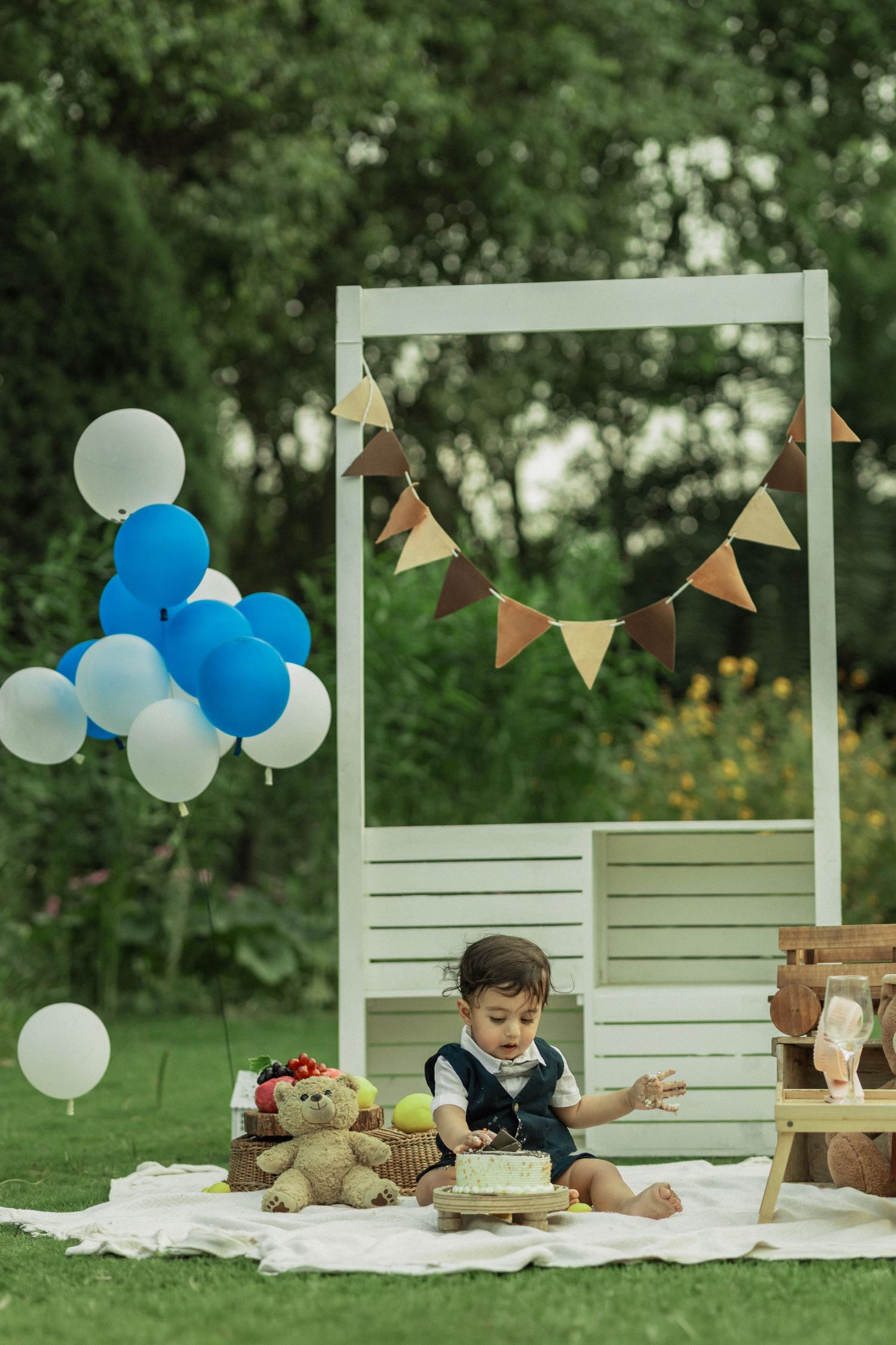 Baby boy celebrates first birthday outdoors with cake, teddy bear, blue balloons, and bunting decorations on a garden blanket.
