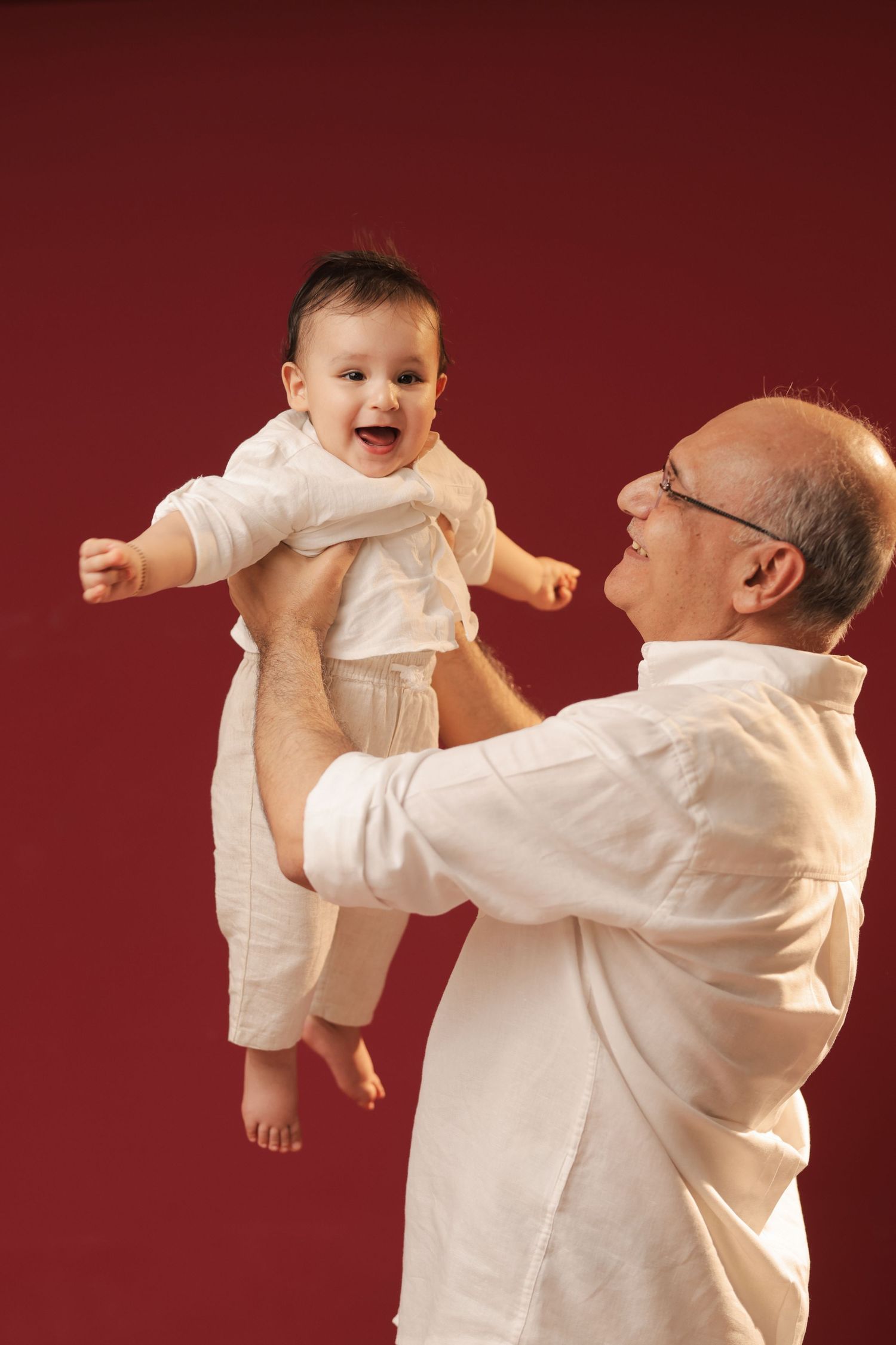 Elderly grandfather in white shirt joyfully lifting laughing baby in white outfit against red background.