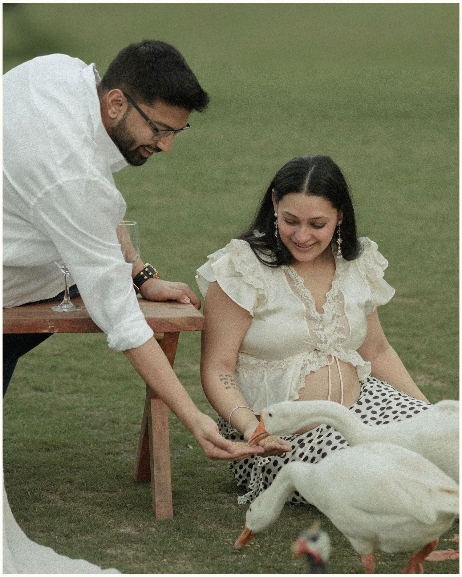 A couple in white outfits feeding ducks on green grass, woman sitting while man leans over a wooden bench.