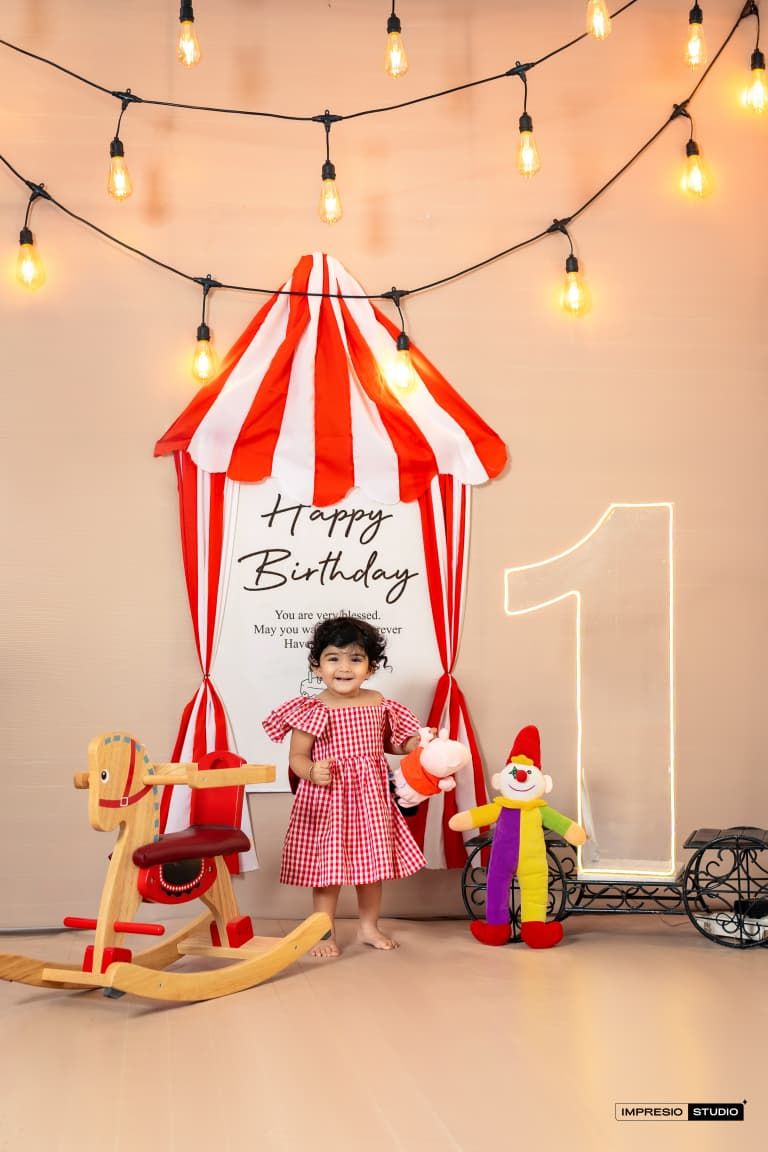 Toddler girl in red gingham dress at circus-themed first birthday party with striped tent, clown, and string lights.