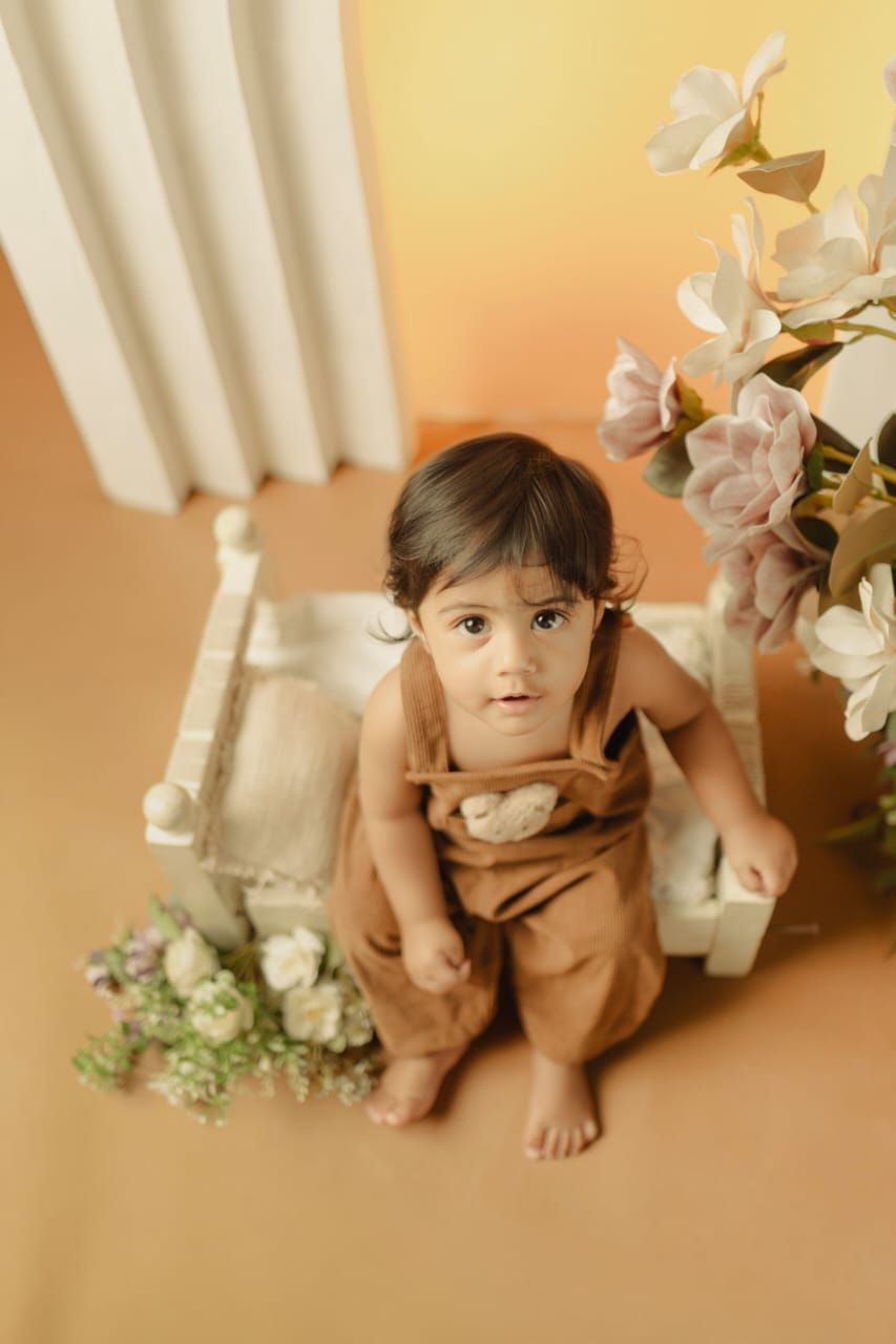 A toddler in a brown romper sits barefoot on a studio floor surrounded by white floral props and a miniature bench.
