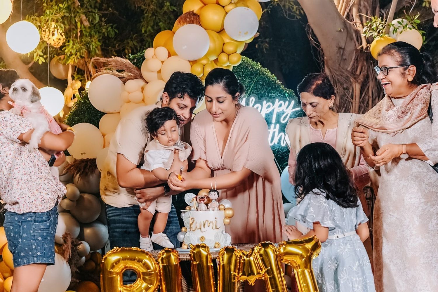 Family gathering around a birthday cake at a festive party with gold balloon decorations and a Happy Birthday backdrop.