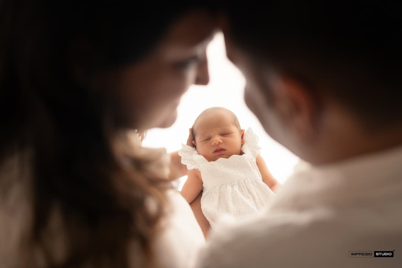 Parents gazing lovingly at their sleeping newborn baby dressed in white, captured in a warm, softly lit portrait.