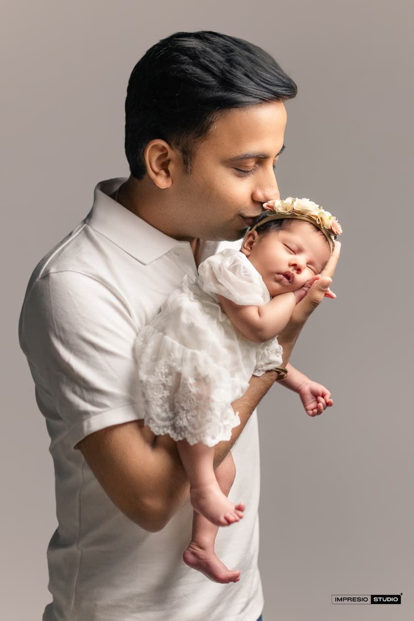 Father in white shirt kissing sleeping newborn baby girl in white dress with floral headband in studio portrait.