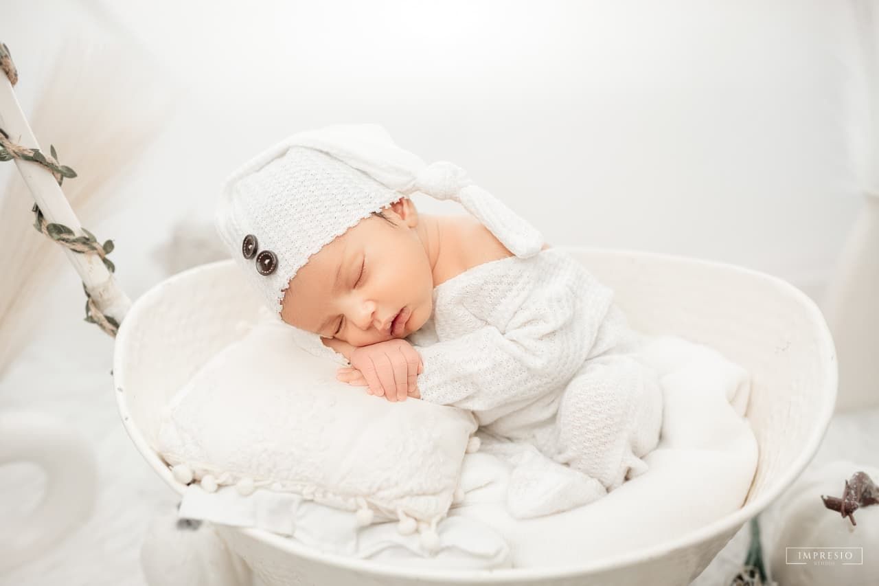 Serene newborn baby in white knit outfit and hat sleeping in a white oval basket with pillow.