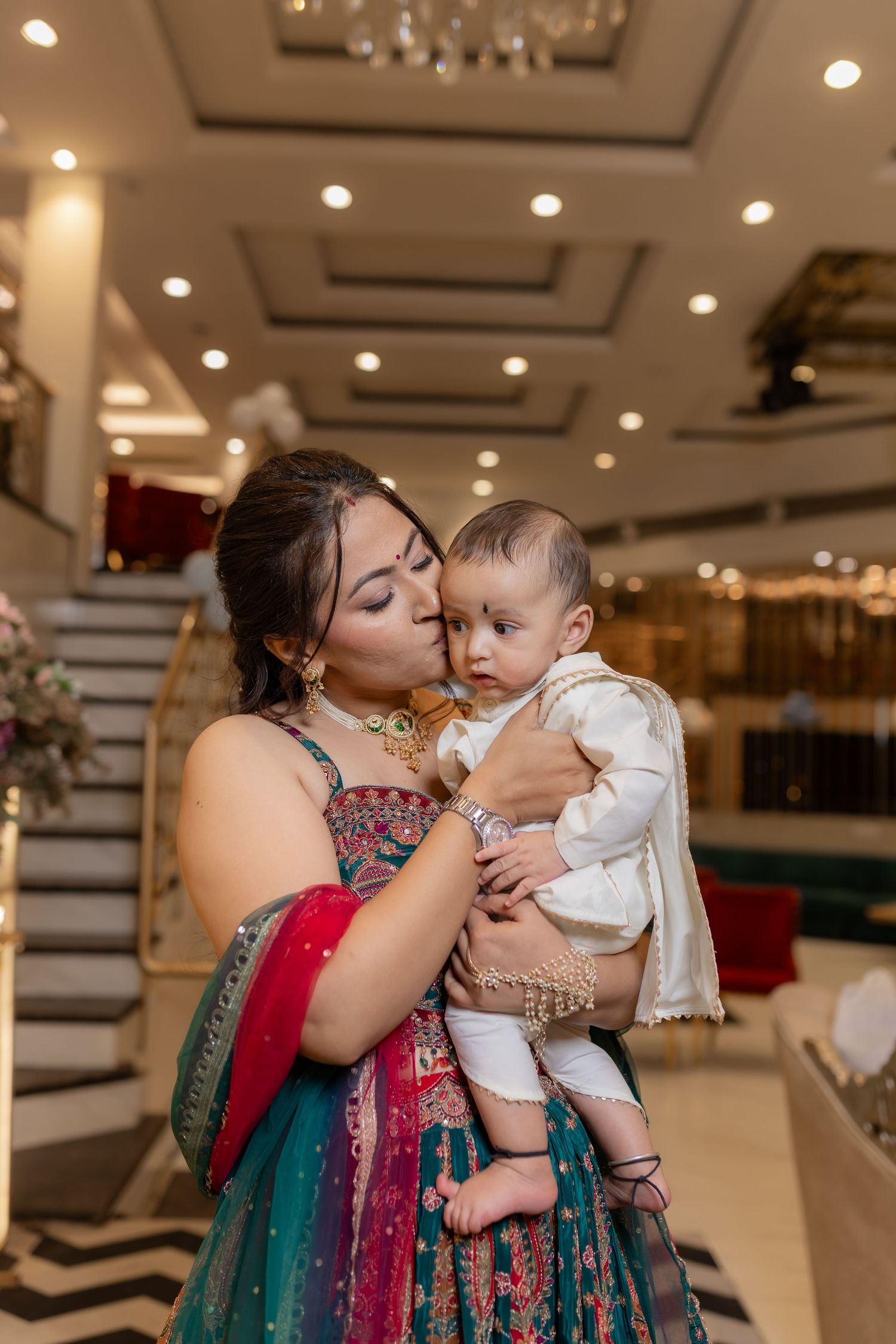 Woman in a teal and red embroidered lehenga kisses a baby in a white outfit inside an elegant banquet hall with a staircase.