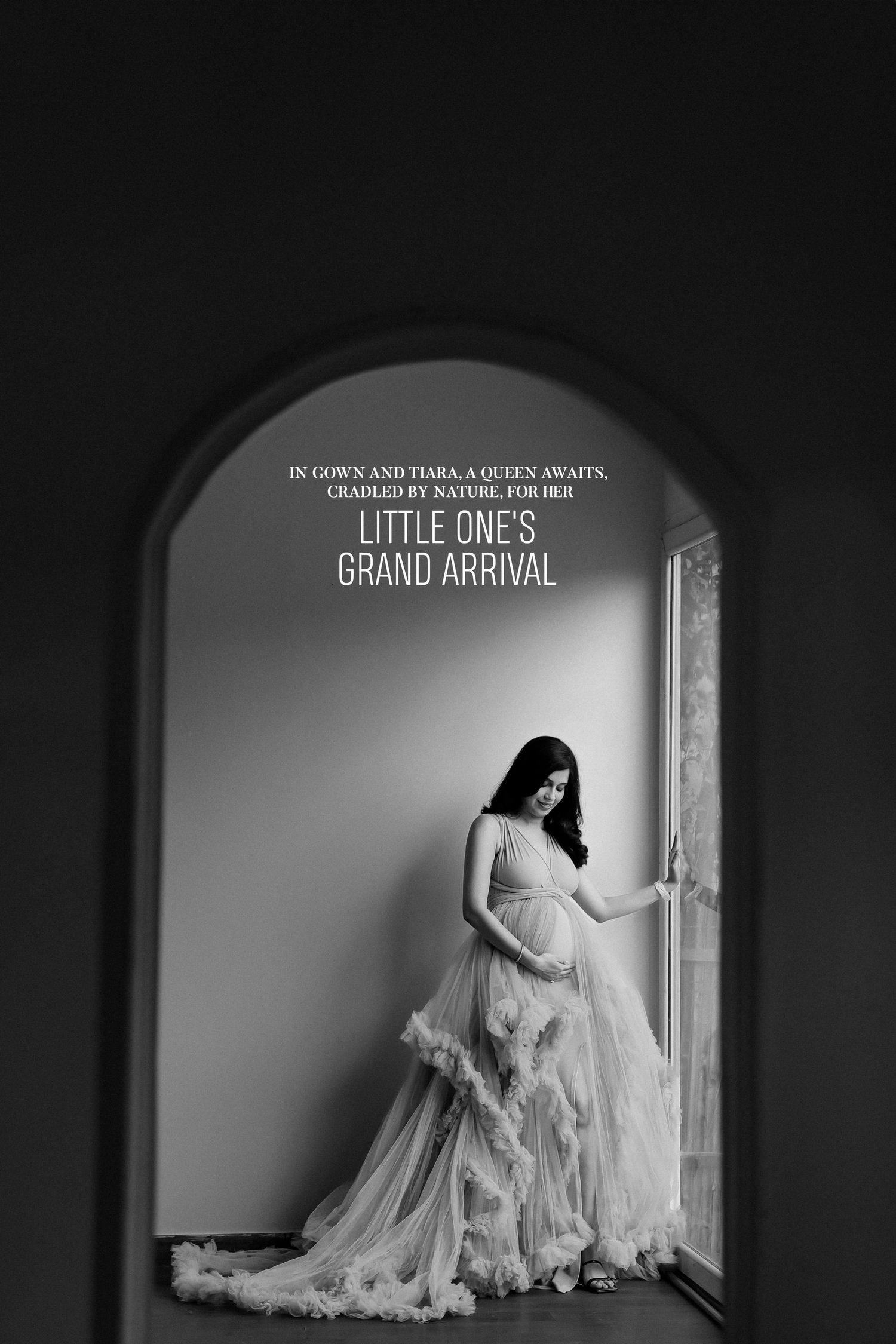 Elegant pregnant woman in flowing tulle gown poses by window in black and white maternity portrait.