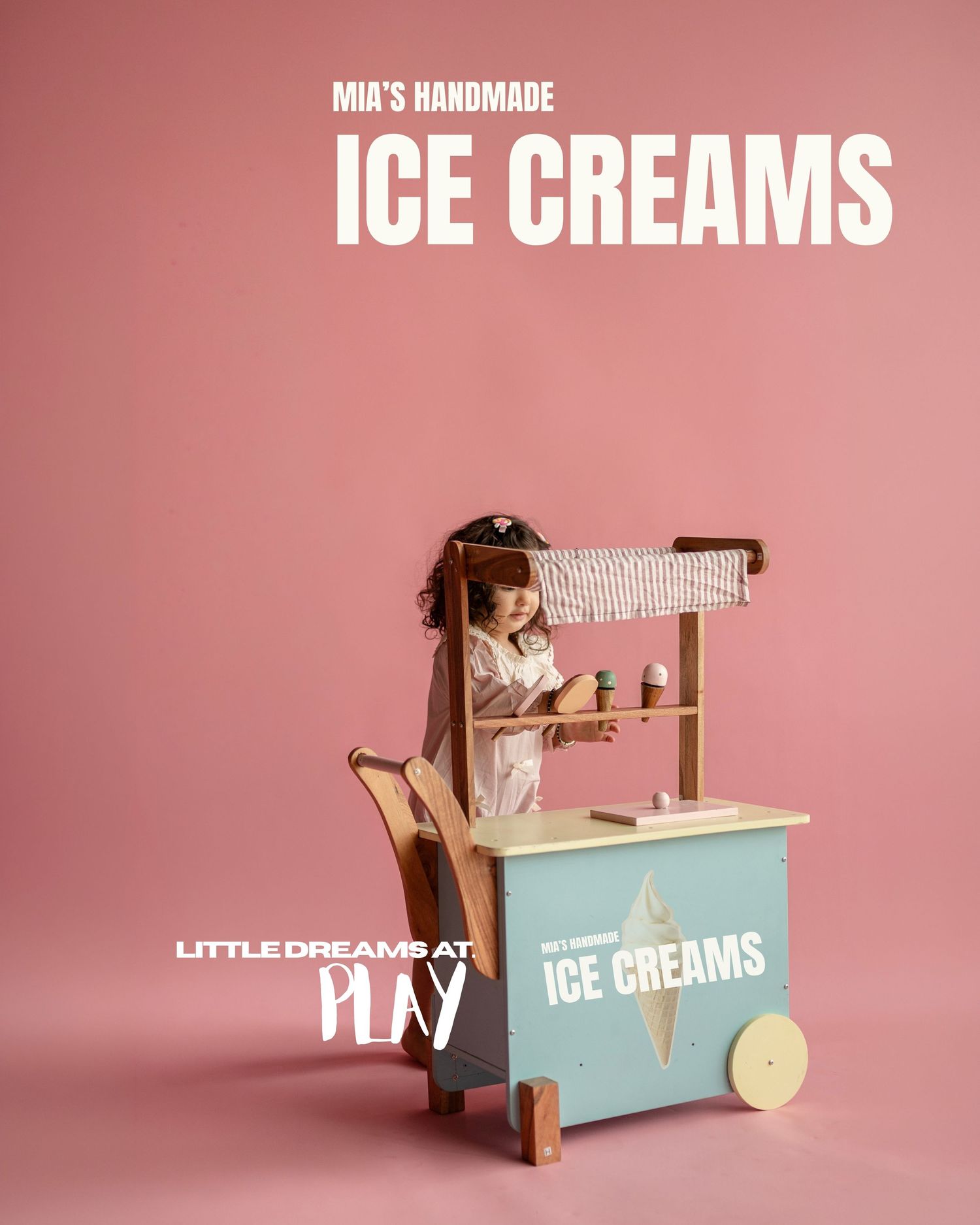 A young girl plays with a wooden toy ice cream cart labeled Mia's Handmade Ice Creams against a pink background.