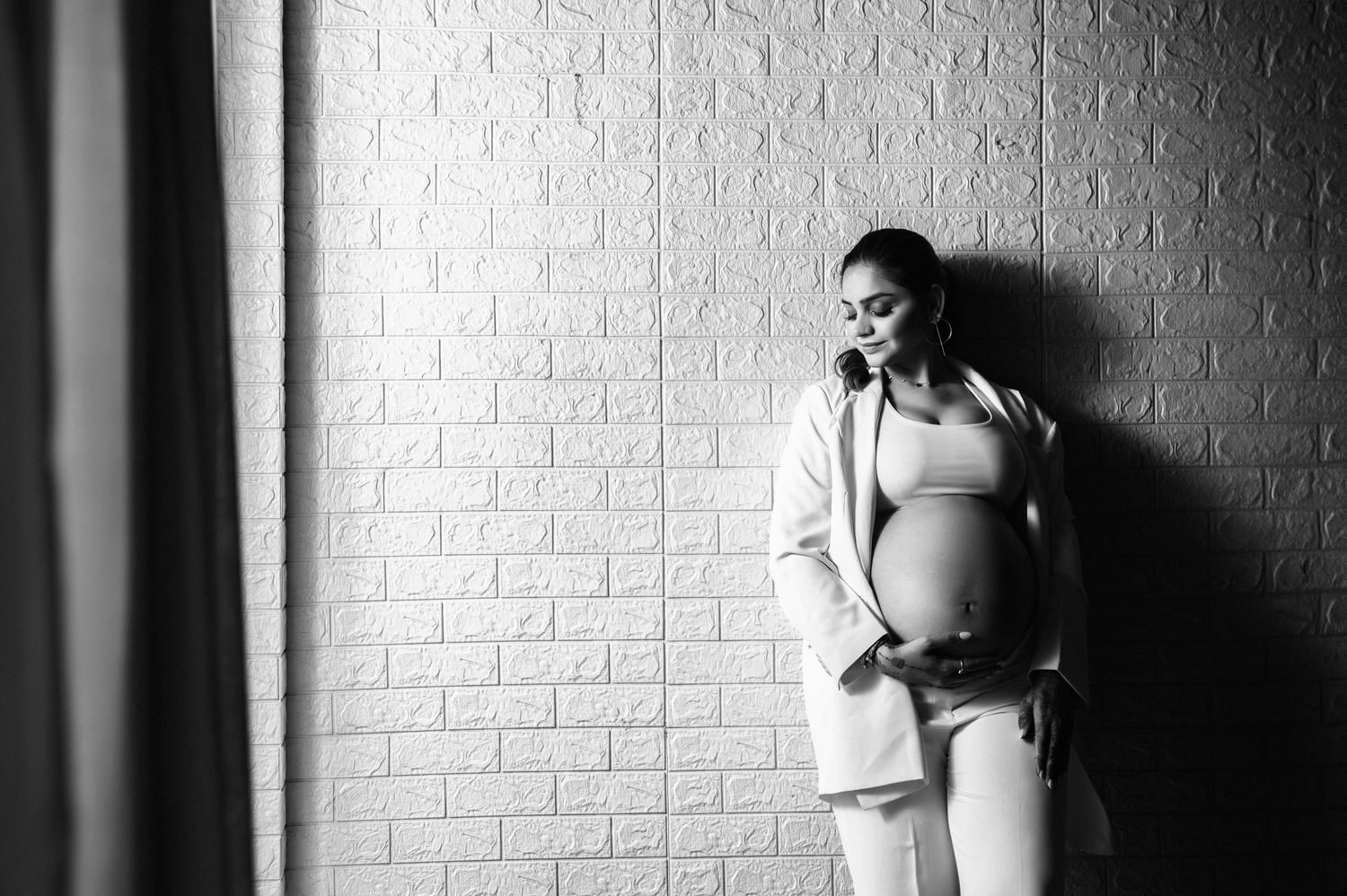 Black and white photo of a pregnant woman in a suit cradling her baby bump against a brick wall.