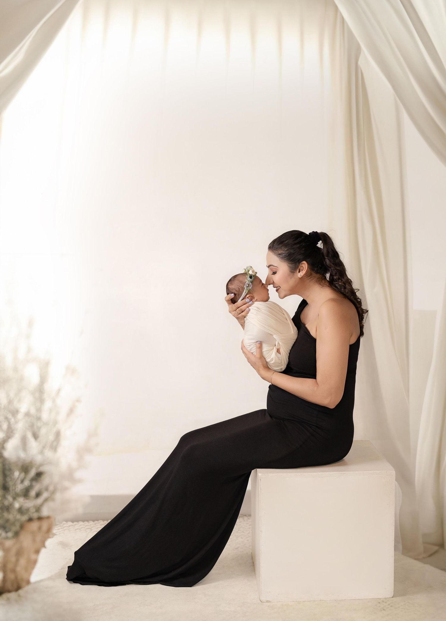 A woman in a black gown sits gracefully, lovingly gazing at her newborn baby in a serene, softly lit studio.