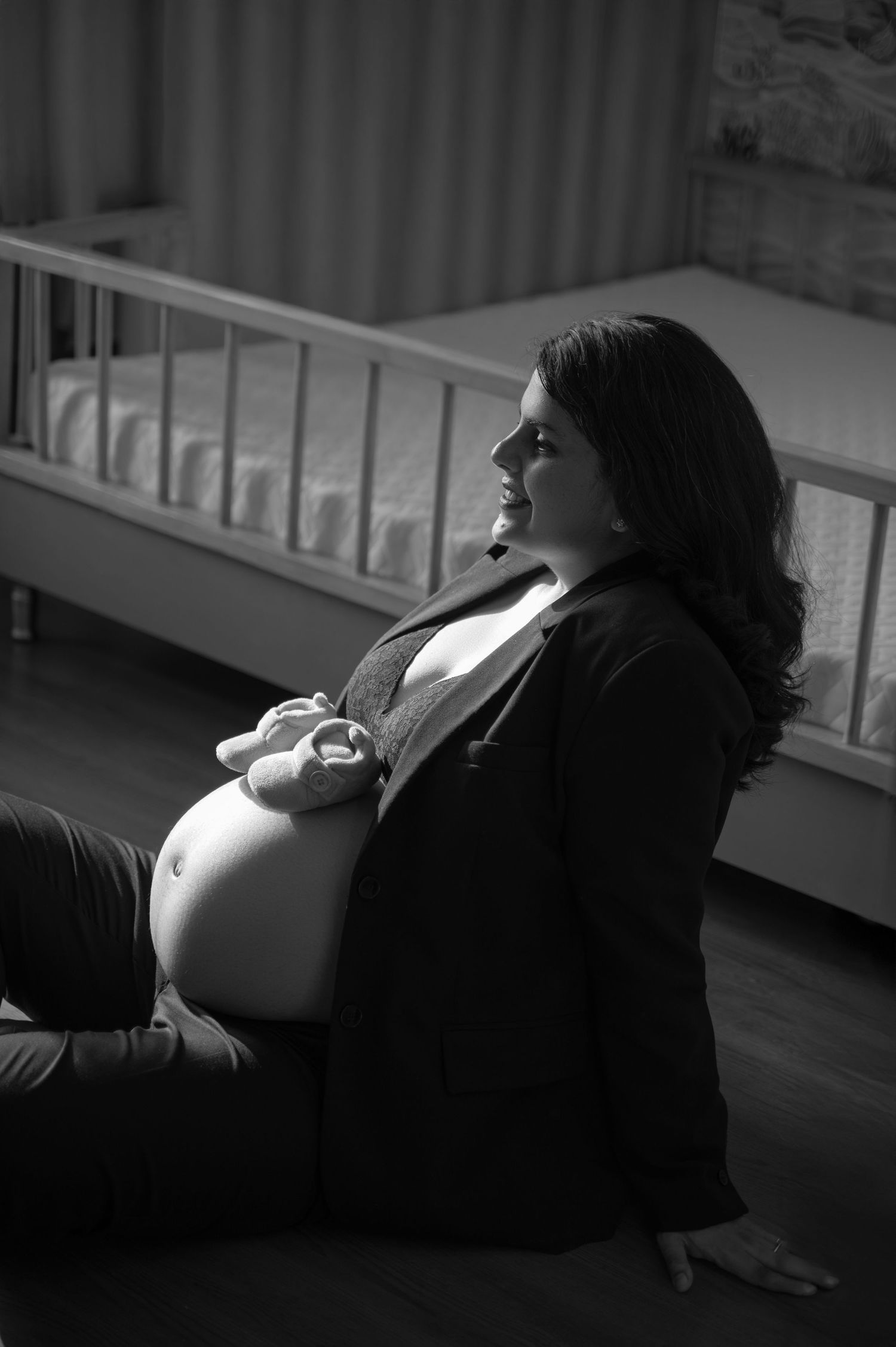 A pregnant woman sits in profile by a crib, captured in dramatic black and white lighting and getting an elegant maternity photoshoot done.