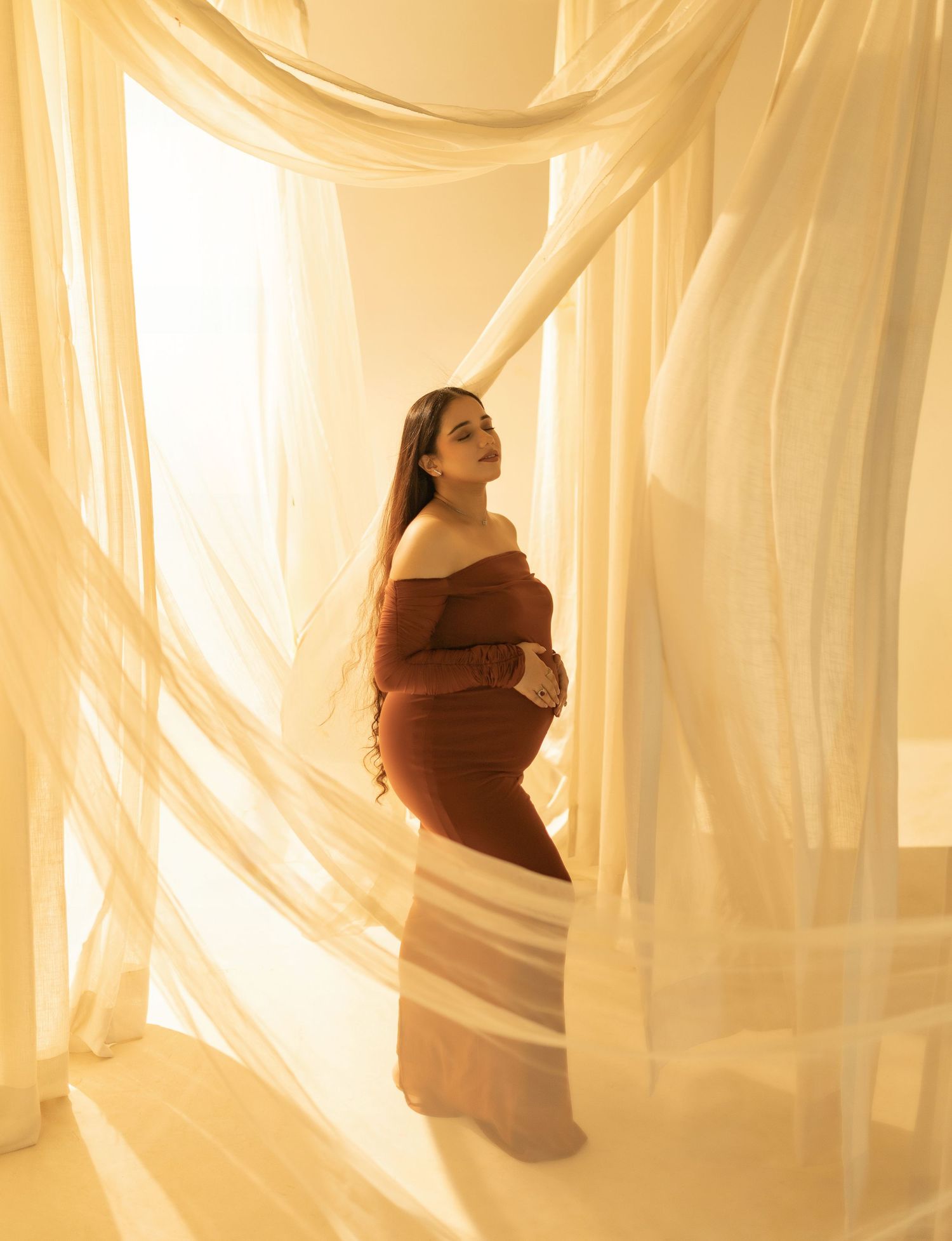 Silhouette of a pregnant figure seen through sheer flowing fabric curtains in warm golden lighting.