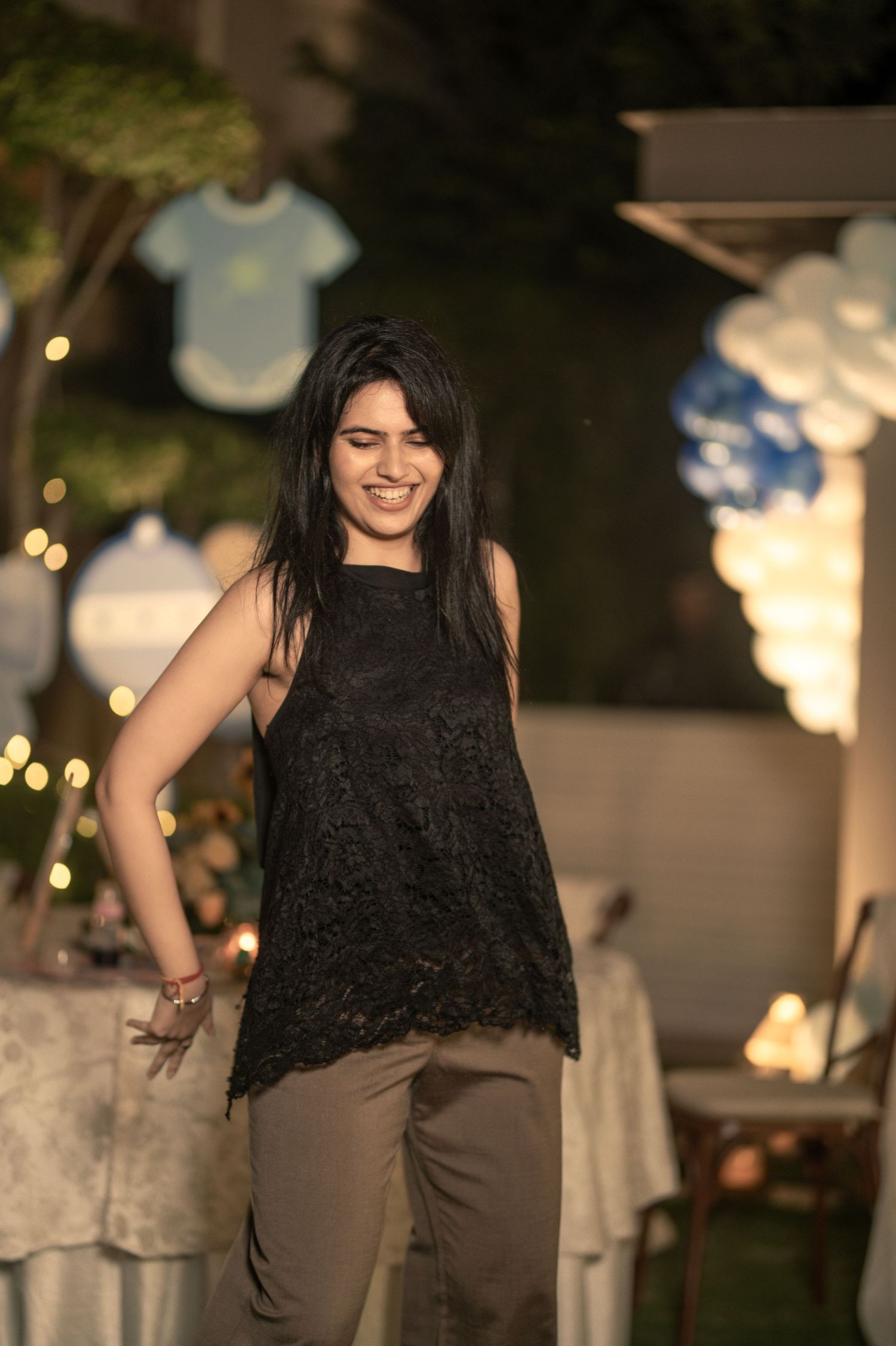 Young woman in black lace top and khaki pants smiling at an outdoor evening party with balloon decorations.