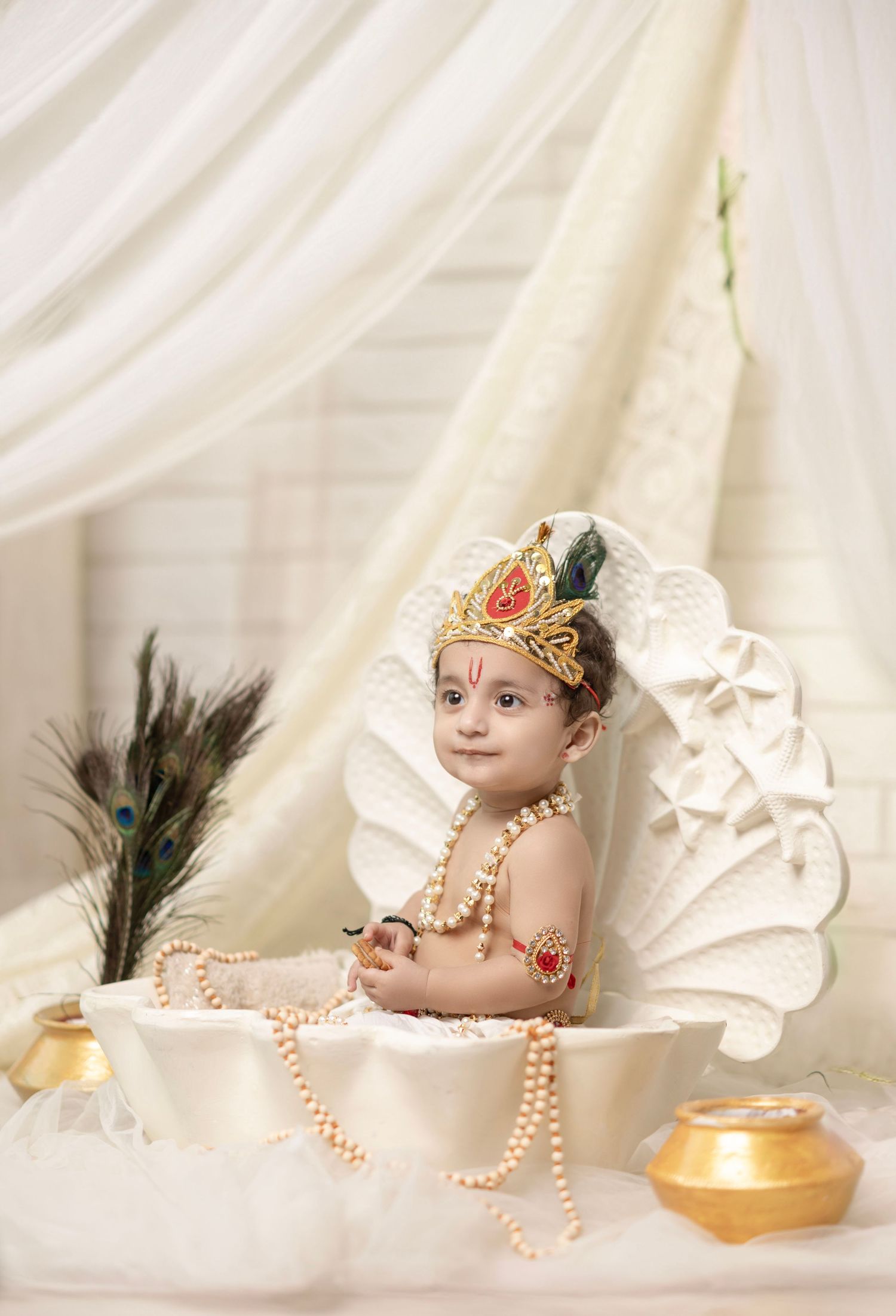 Baby dressed as Lord Krishna with crown, pearl necklaces, and traditional jewelry, seated in decorative shell prop.