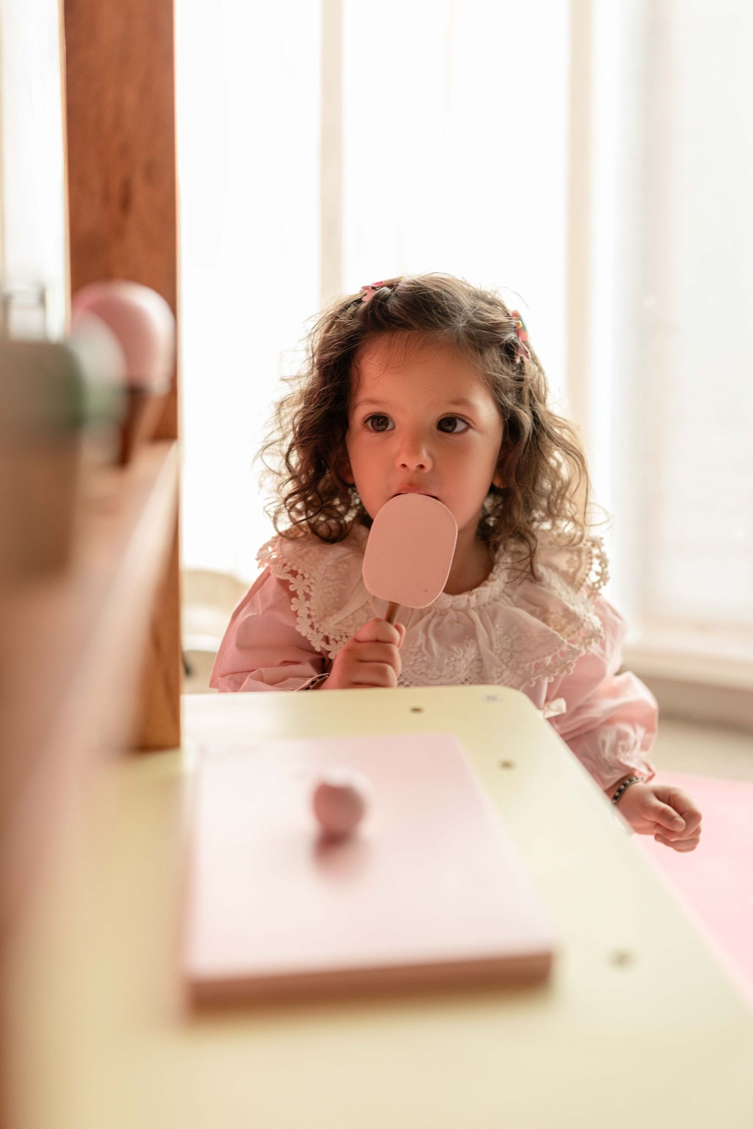 Young girl in pink dress eating a pink popsicle at a white table indoors.