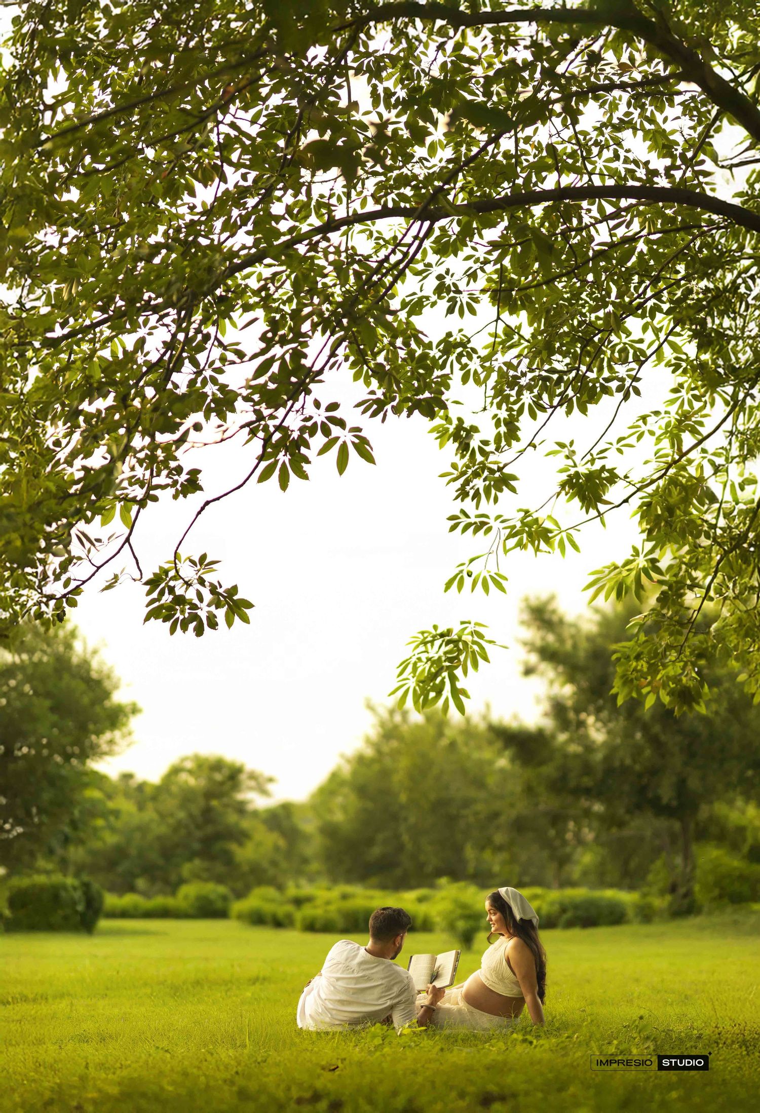 Couple sitting on lush green grass under a large tree in a serene park setting.