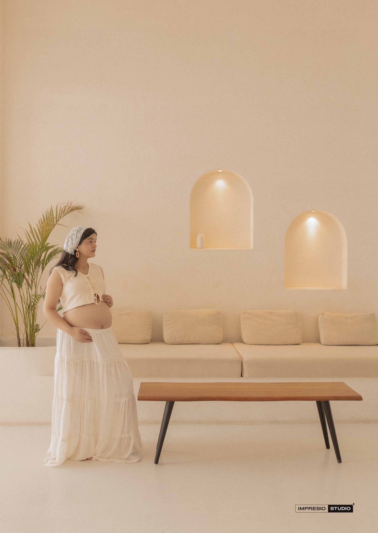 Pregnant woman in white dress poses in minimalist beige studio with arched wall niches and wooden coffee table.